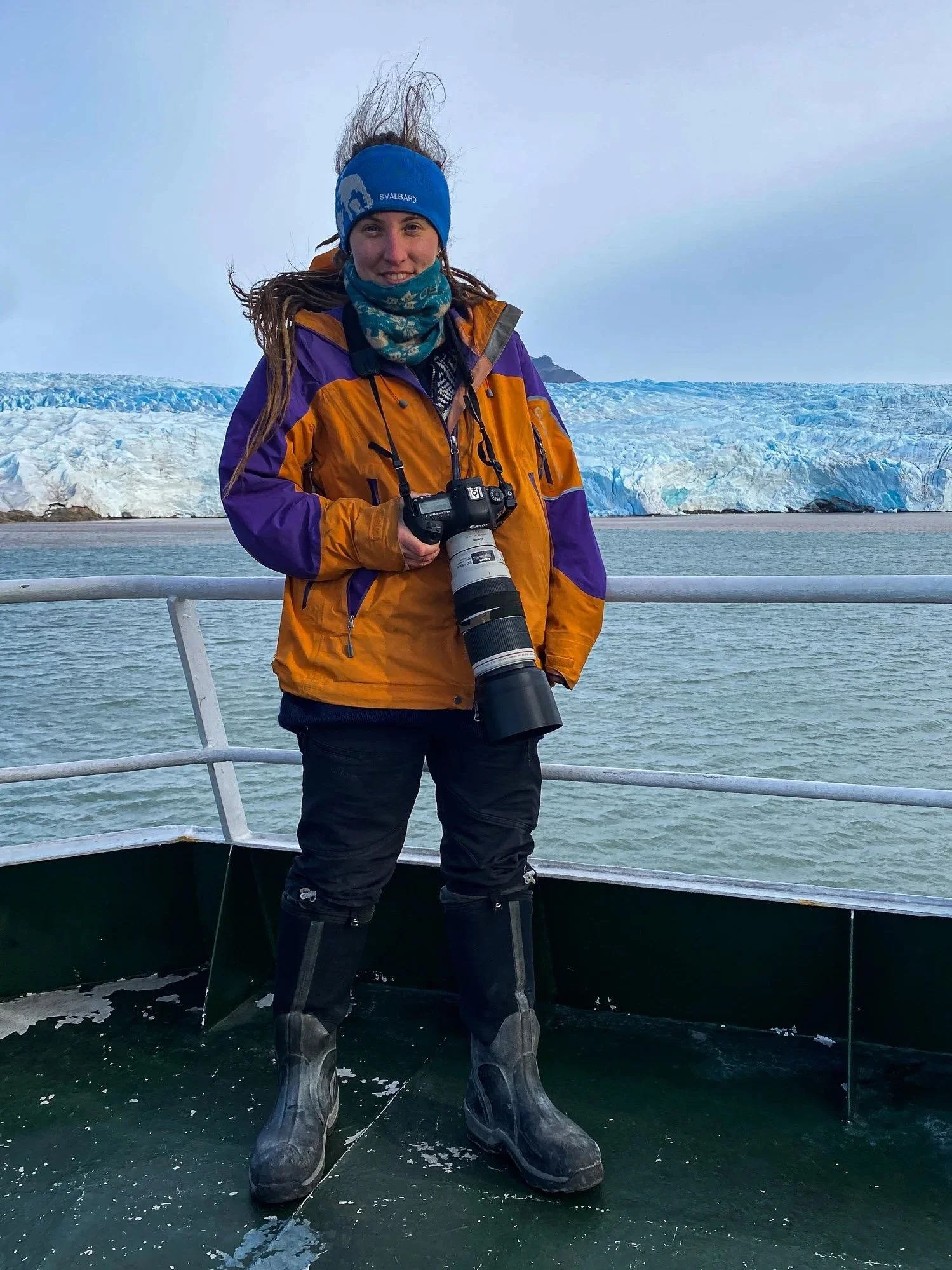 A woman (Laia) in outdoor gear holding a camera with a long lens on a boat, with glaciers and water in the background.