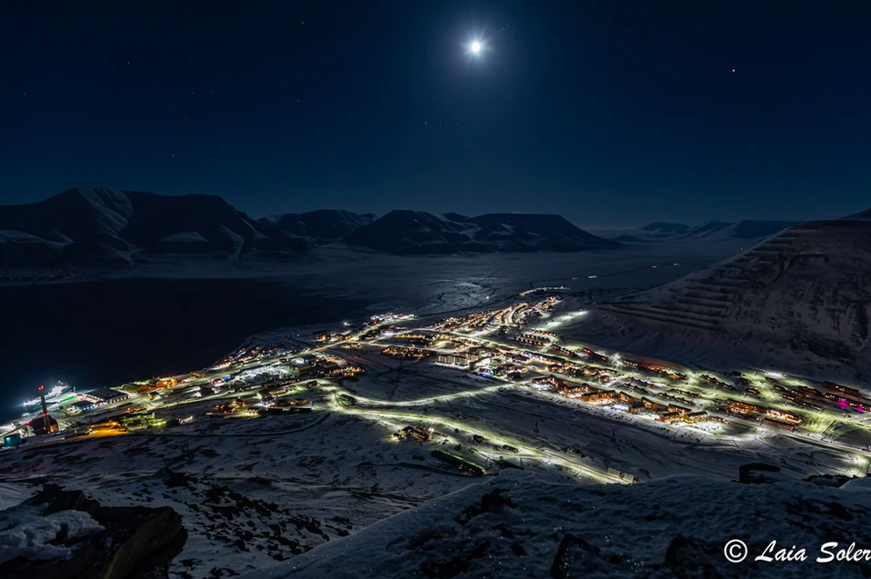 Nighttime view of a snow-covered mountain village illuminated by streetlights under a bright moon and starry sky.