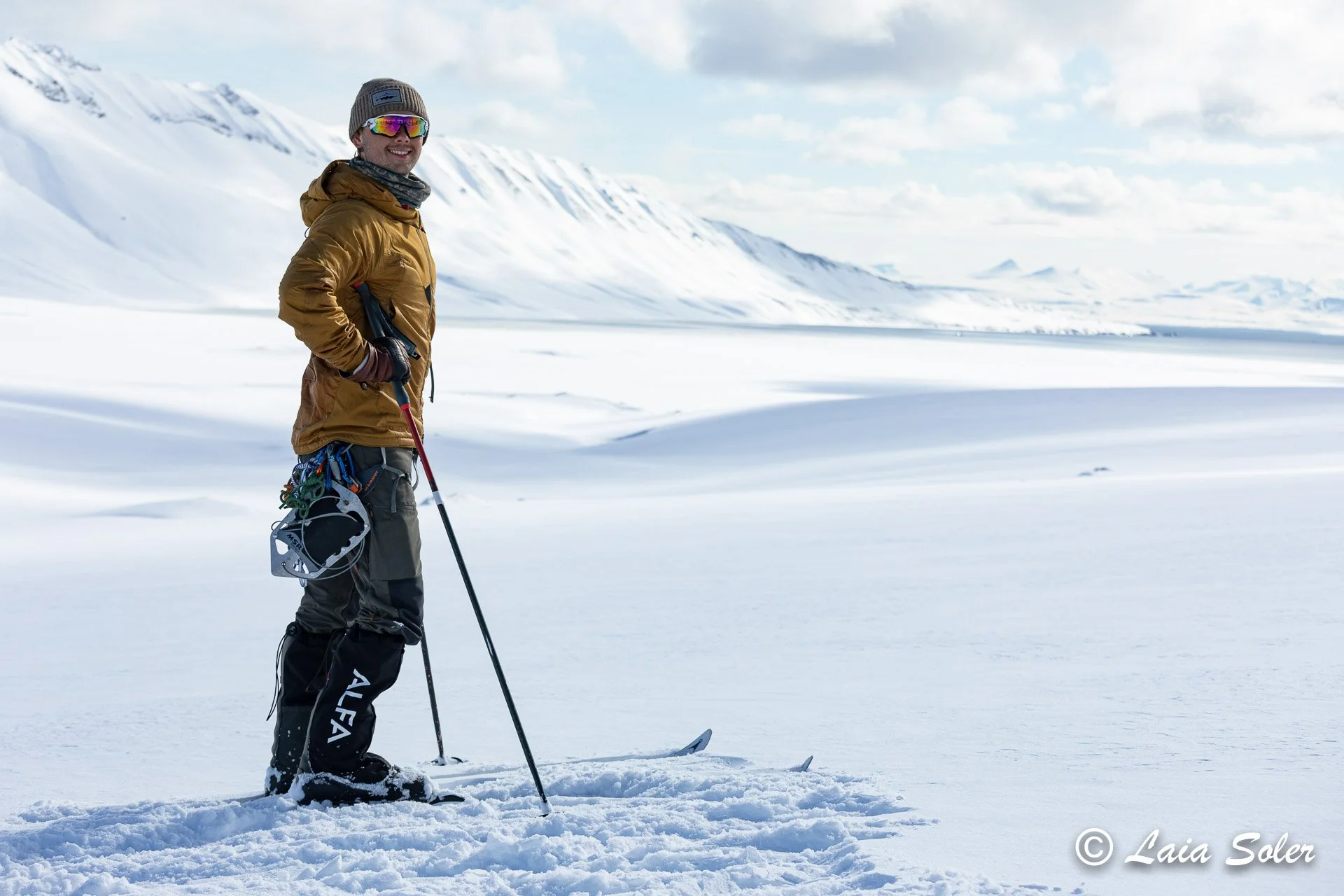 A man stands on skis on a snow-covered glacier with mountains in the background, dressed in winter gear, holding ski poles, and smiling at the camera in a snowy landscape.