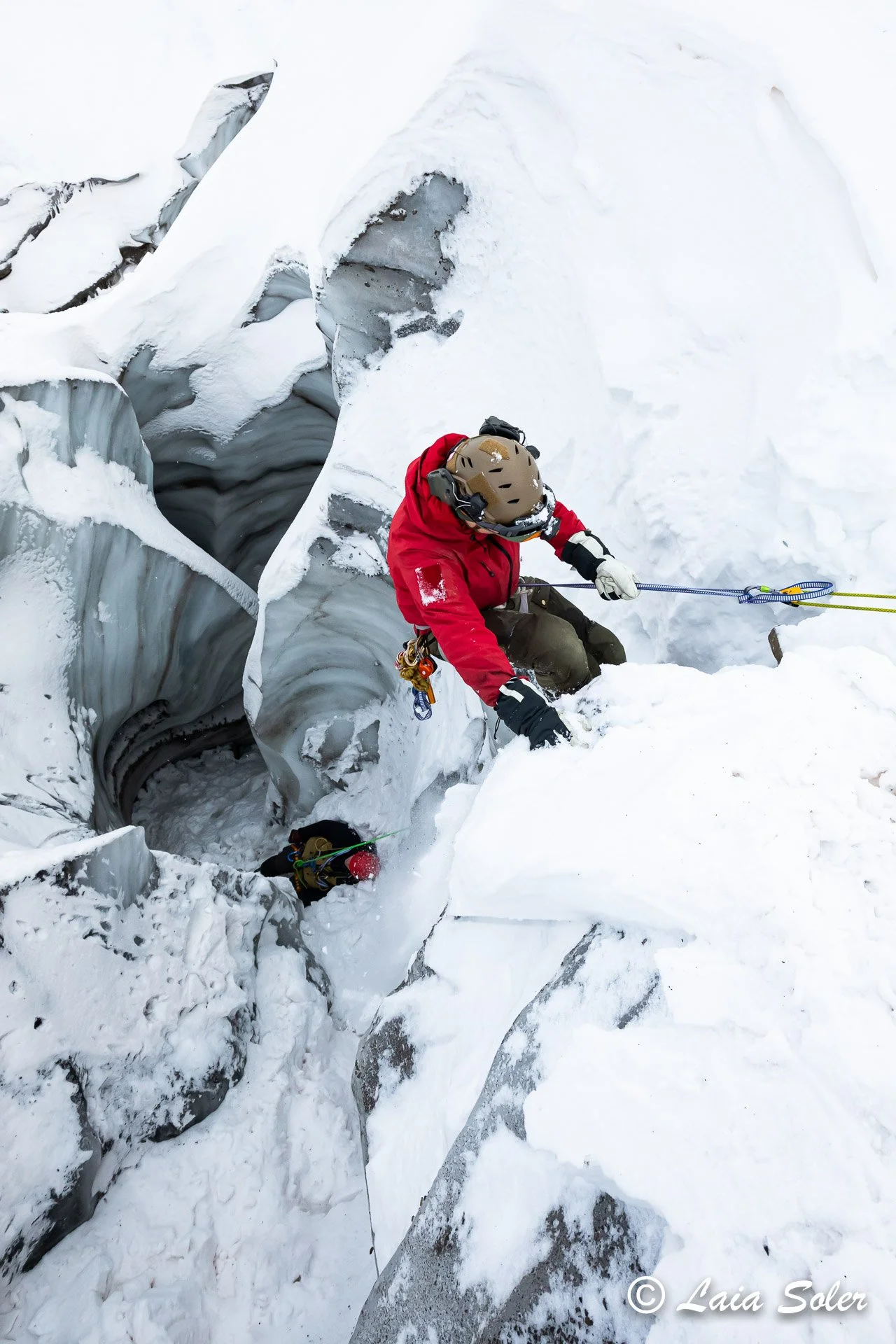 A person in a red jacket and climbing gear is descending into a meltwater channel to rescue an unconscious person on a snow-covered glacier.