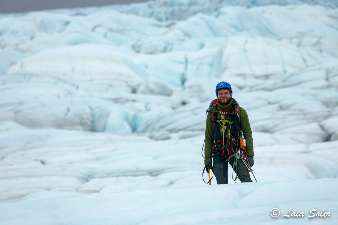 A man in climbing gear standing on ice in a glacier with blue and white ice formations in the background.