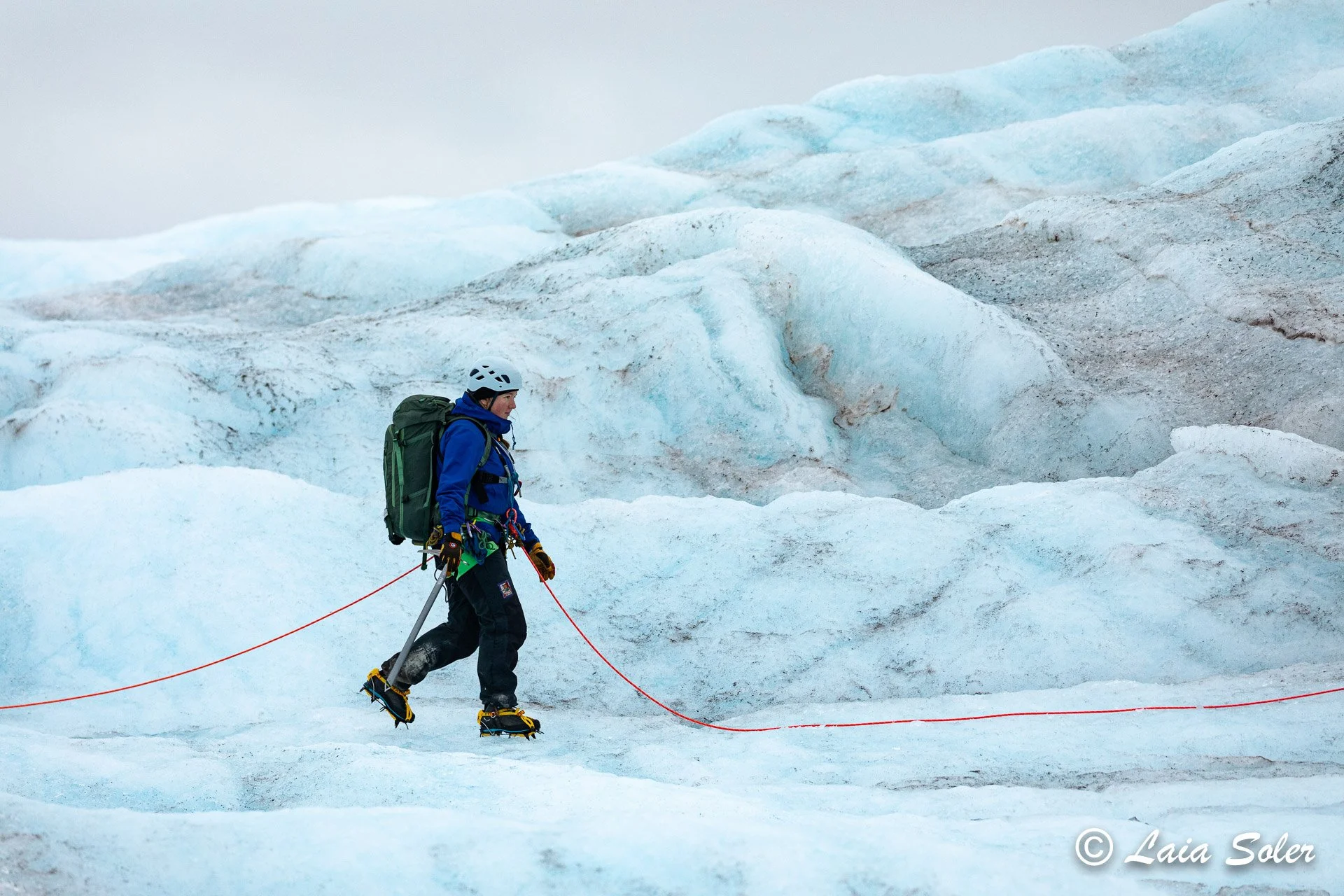 A person in winter gear, including a helmet, blue jacket, backpack, and crampons, walking on snow and ice near large ice formations, in a cold, icy environment.