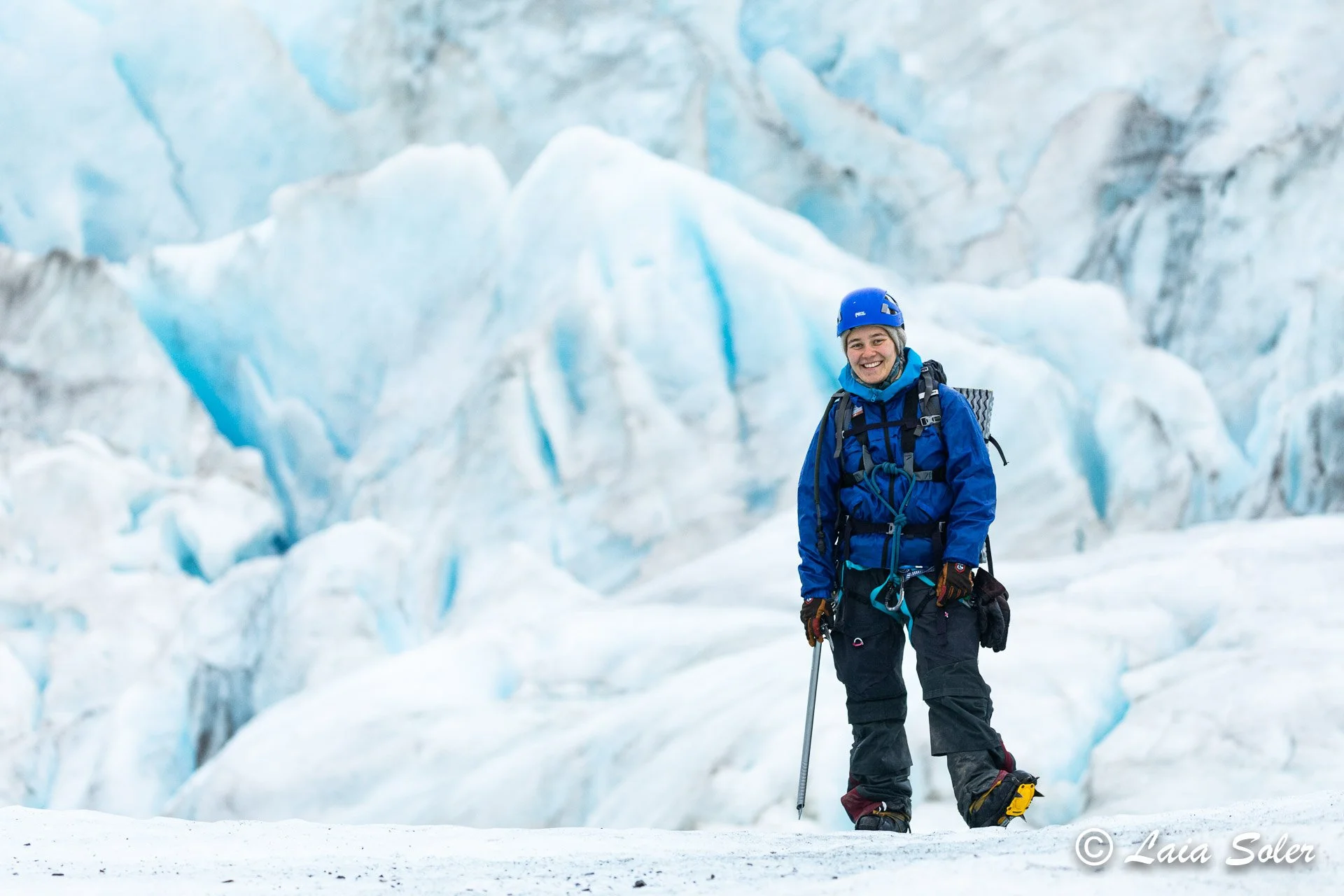 A person in winter gear, including a blue helmet and jacket, standing on the glacier with crevasses in the background, smiling at the camera.