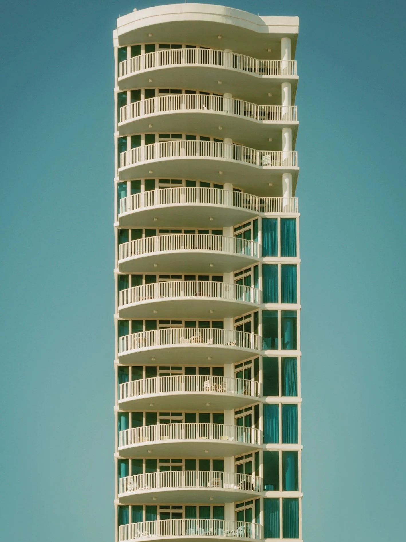 A minimalist modern apartment building with multiple curved balconies, large glass windows with blue curtains, and a clear blue sky in the background.