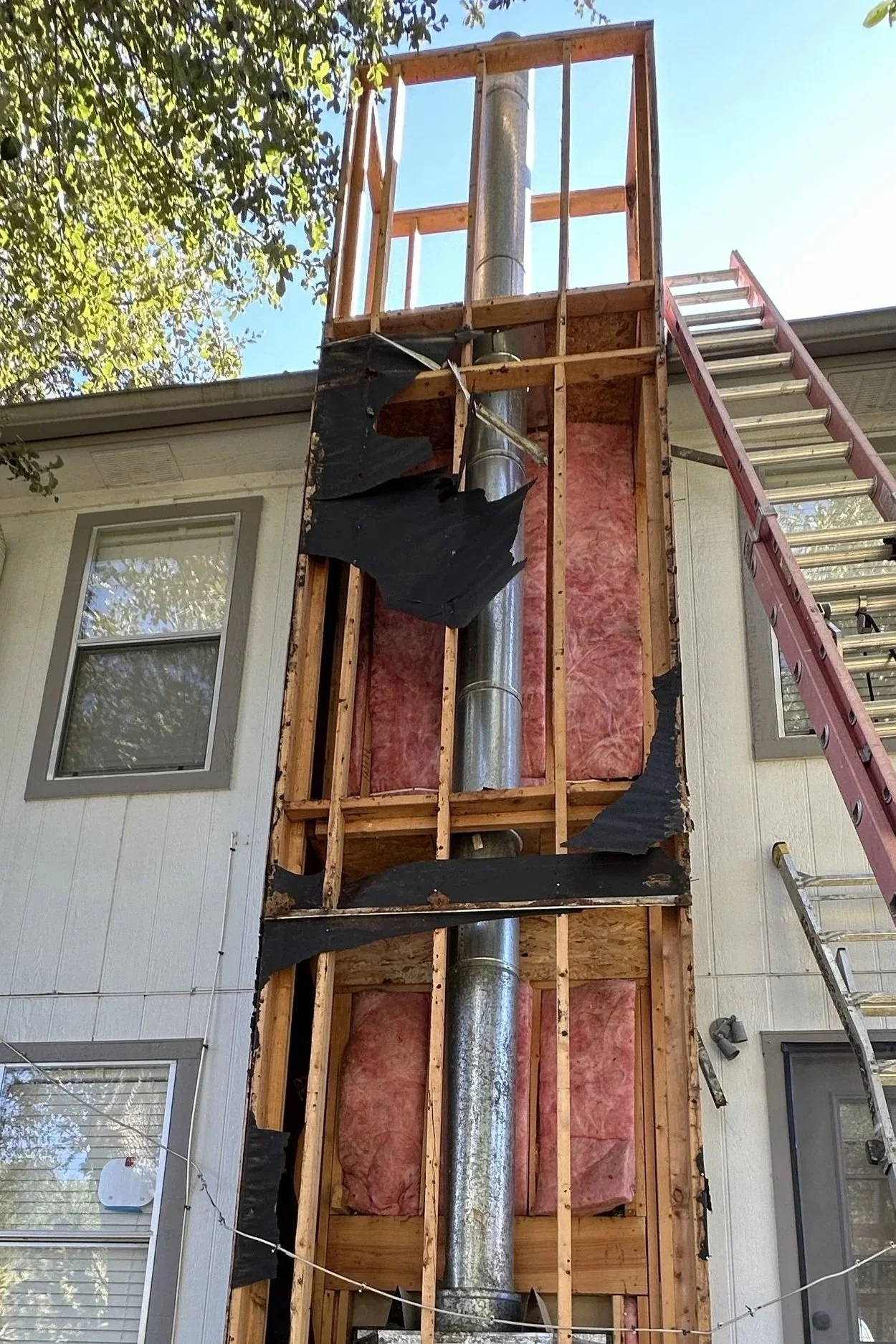 Construction work on the exterior of a house, showing the wall with missing siding and insulation, a metal chimney, and ladder leaning against the wall under a tree.