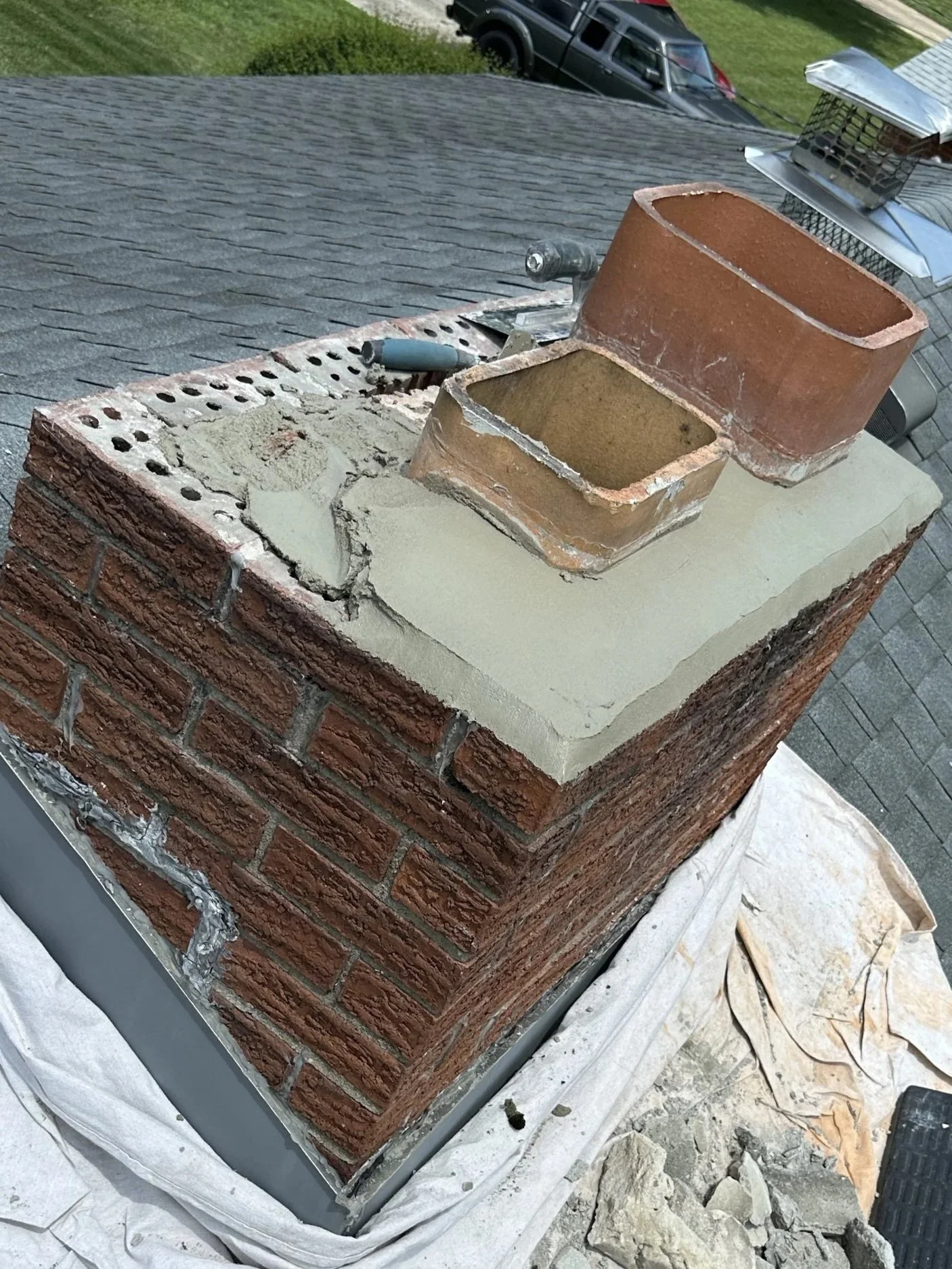 Close-up of a chimney on a house roof, showing the brick structure and a new layer of cement around the chimney base, with some tools nearby.