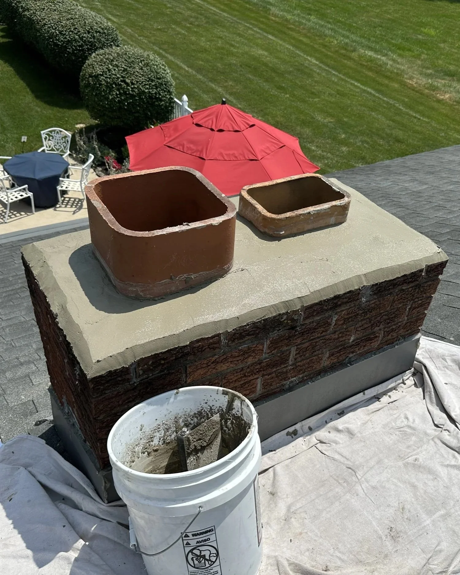 A rooftop chimney repair in progress with two new bricks placed on top, a bucket of mortar nearby, and a tarp covering part of the roof. In the background, there's a yard with outdoor furniture and a red patio umbrella.