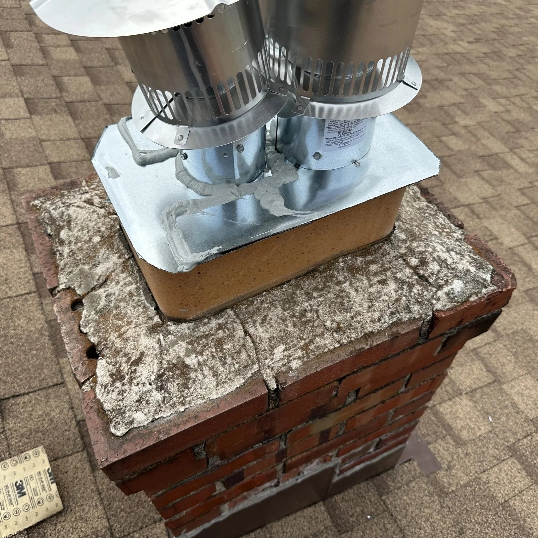 Close-up of a chimney cap mounted on a brick chimney, with roofing and construction materials nearby.