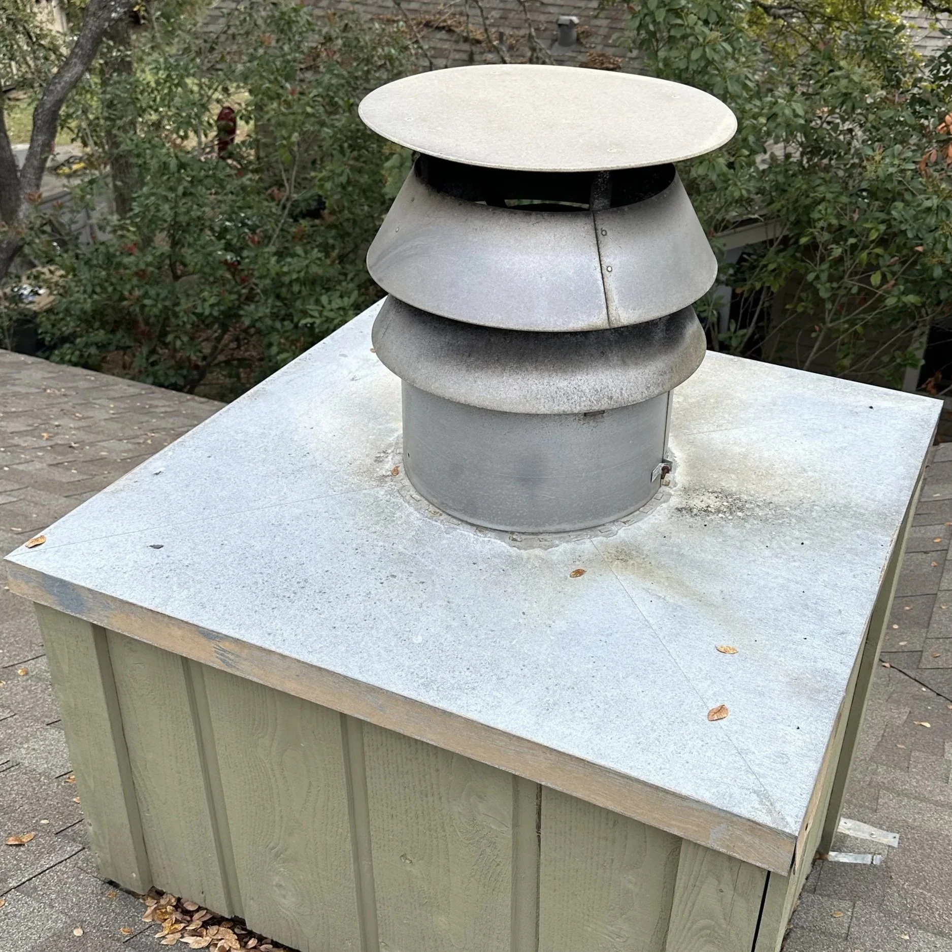 Metal roof vent on top of a small wooden structure with shingles and trees in the background.