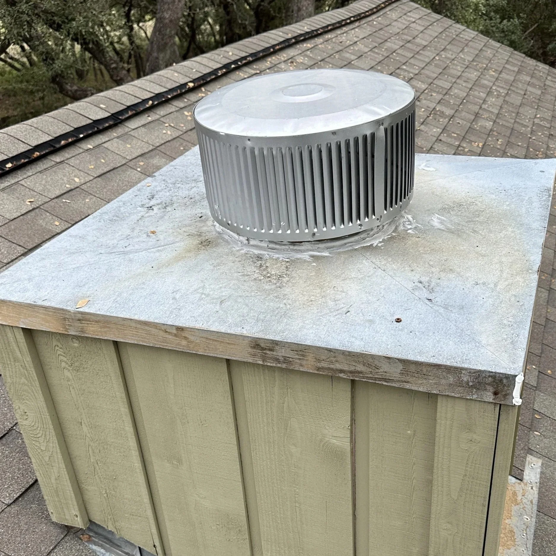 View of a roof vent on a chimney with shingle roof and trees in the background.