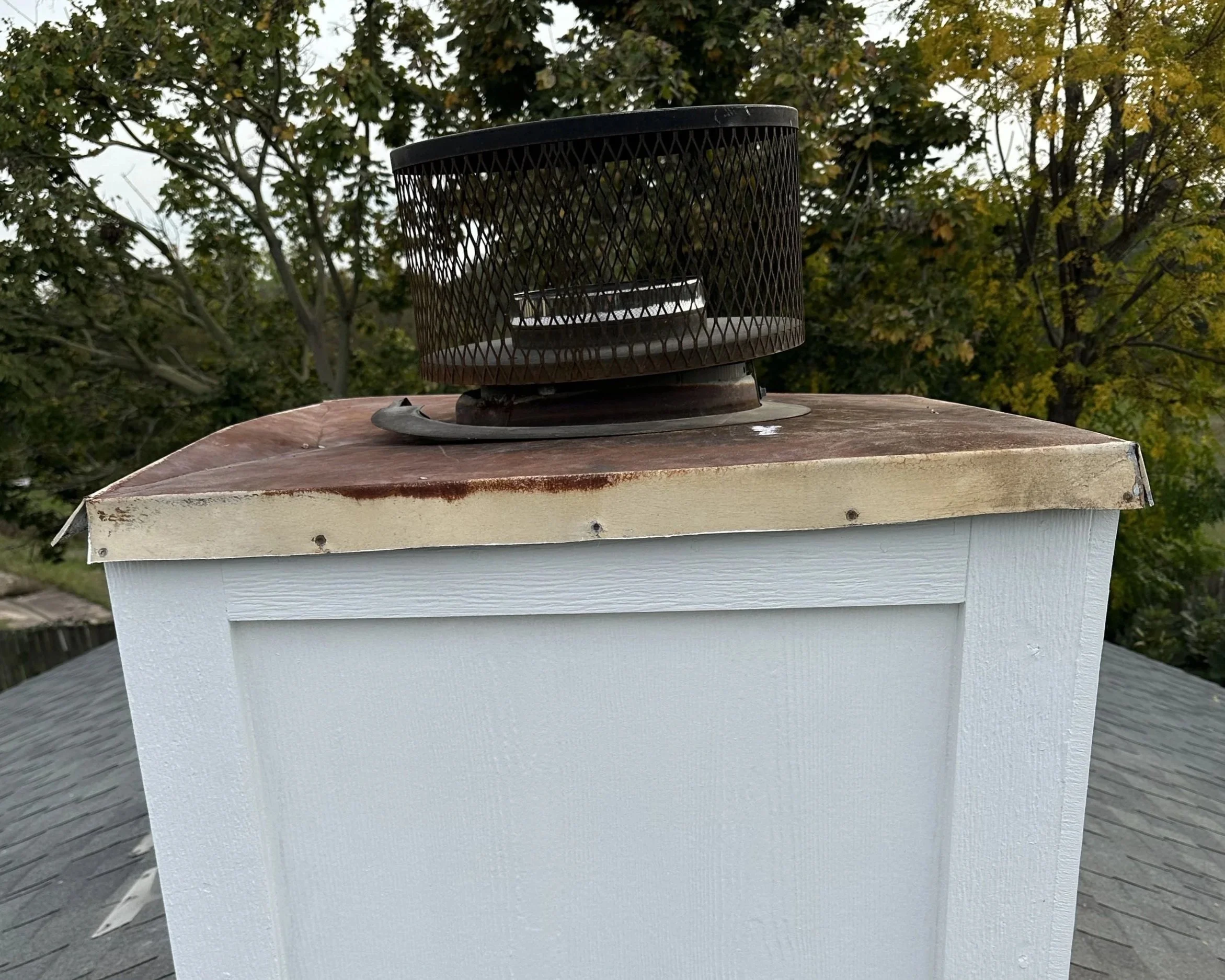 Close-up of a white building exterior with a rusted metal roof vent from a chimney, with background of green trees and a cloudy sky.