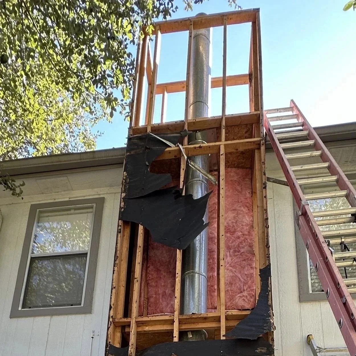 Construction work on the exterior of a house, showing the wall with missing siding and insulation, a metal chimney, and ladder leaning against the wall under a tree.