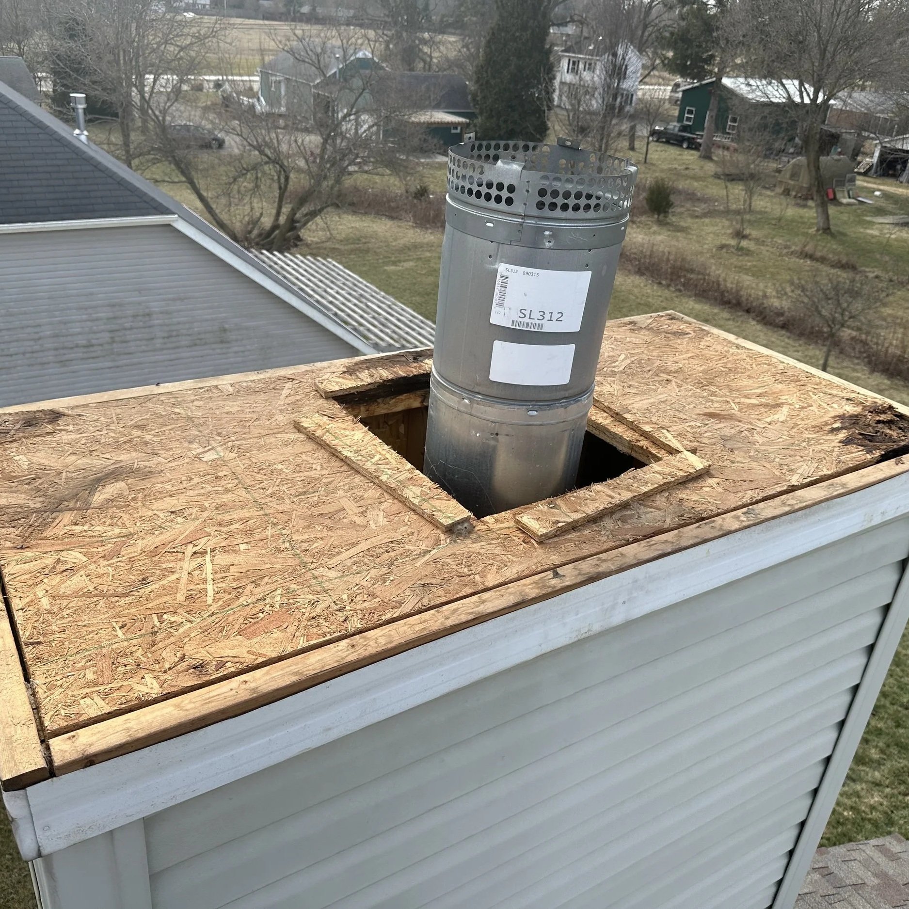 The image shows a wooden roof vent cap on top of a white house with siding, with a metal vent pipe protruding through a square opening in the wooden roof surface. The roof surface is unfinished OSB plywood.