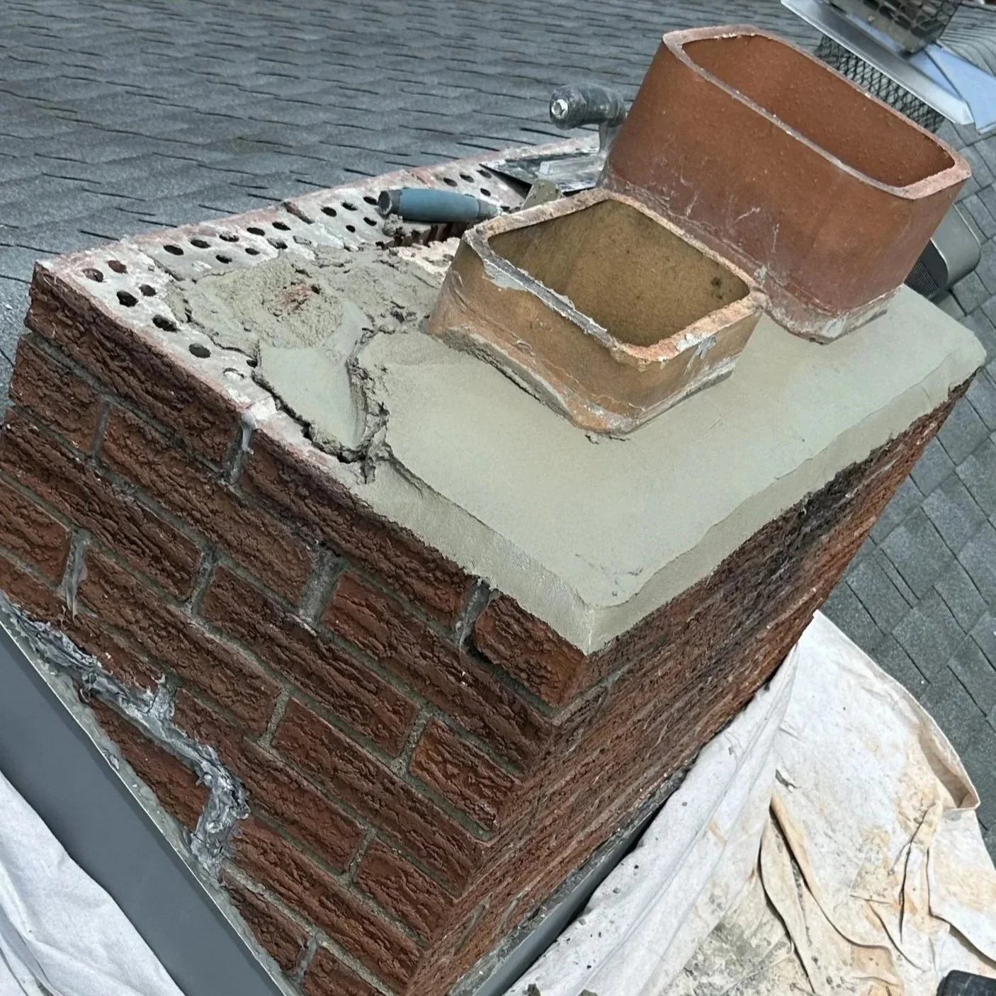 Close-up of a chimney on a house roof, showing the brick structure and a new layer of cement around the chimney base, with some tools nearby.