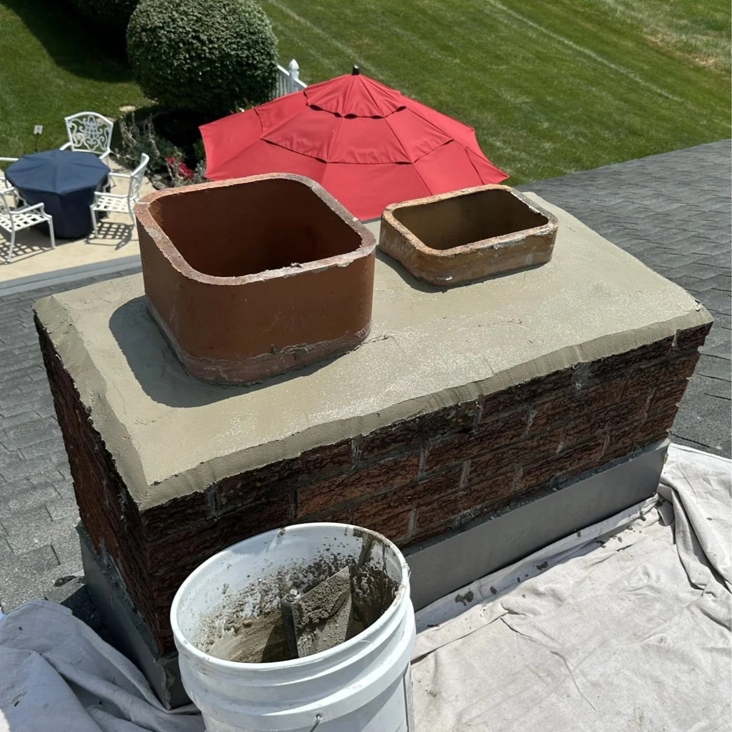 A rooftop chimney repair in progress with two new bricks placed on top, a bucket of mortar nearby, and a tarp covering part of the roof. In the background, there's a yard with outdoor furniture and a red patio umbrella.