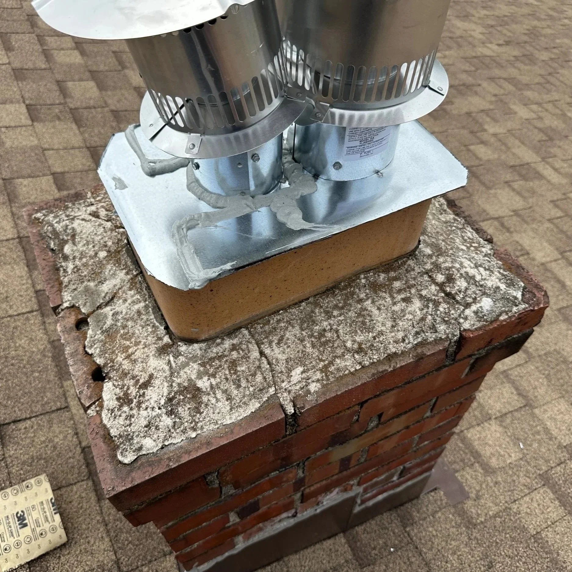 Close-up of a chimney cap mounted on a brick chimney, with roofing and construction materials nearby.
