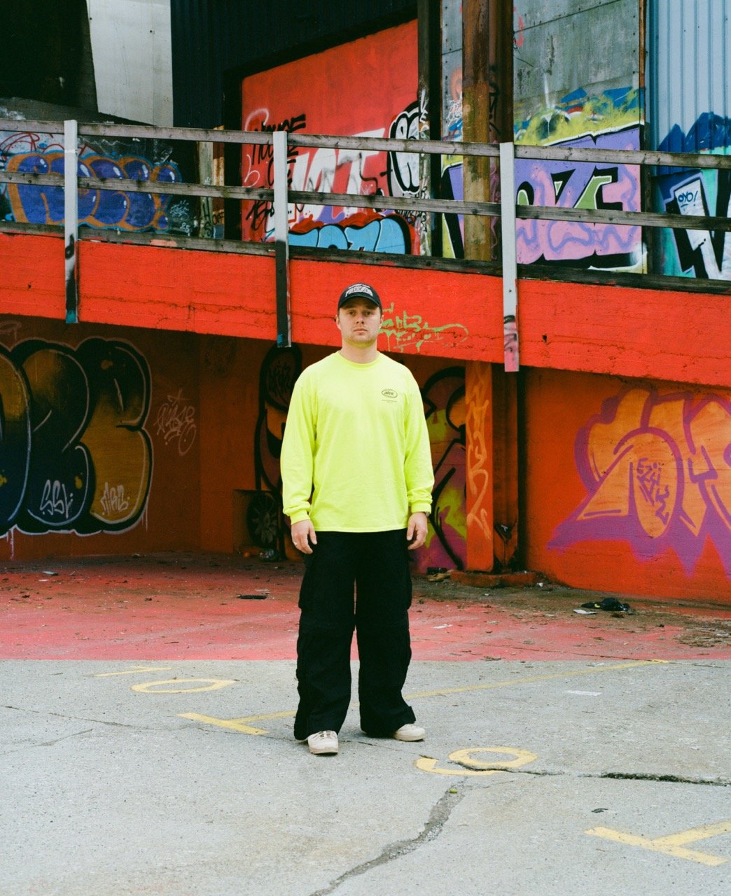 A young man wearing a neon yellow long-sleeve shirt, black pants, and a black cap stands in front of a graffiti-covered wall and colorful urban backdrop.