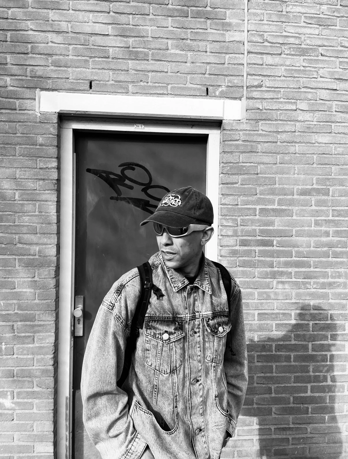 A man wearing sunglasses, a baseball cap, and a denim jacket stands against a brick wall with a door behind him.