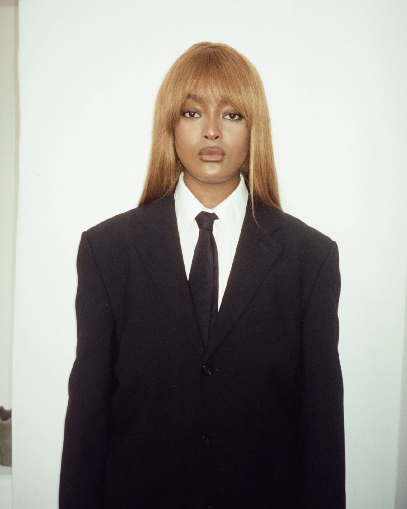 A woman standing in front of a plain white background, dressed in a black suit with a white shirt and black tie.