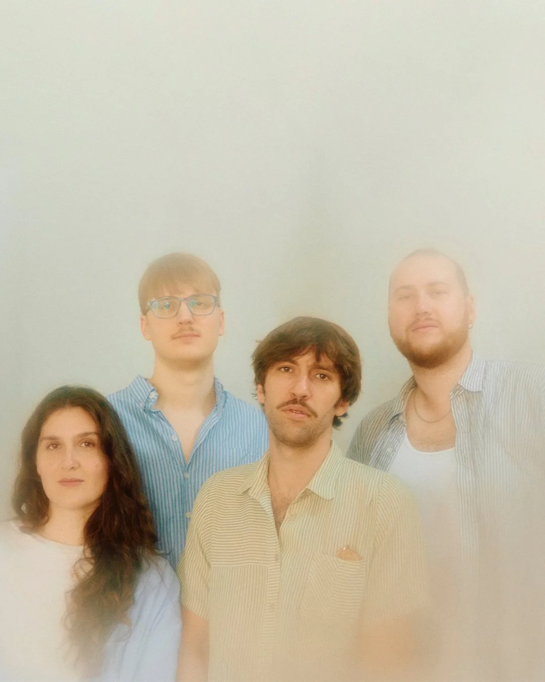 A group of four young adults posing for a photo against a pale background, with one woman and three men, all with casual clothing and relaxed expressions.