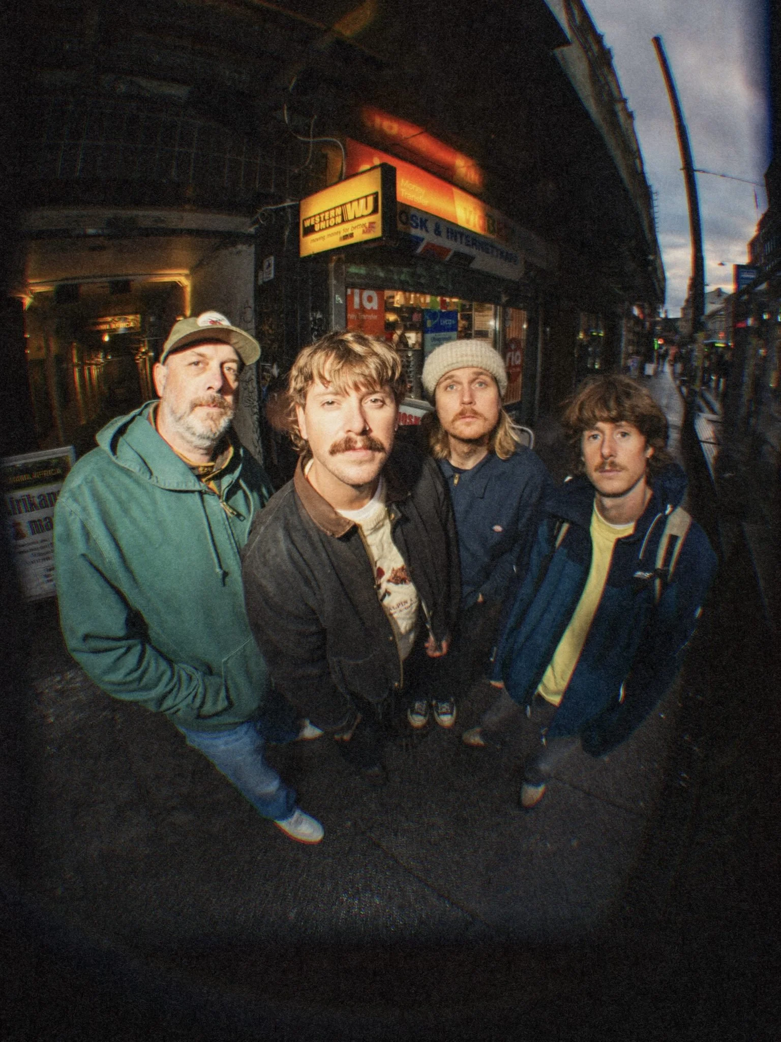 Four men standing on a city street at night, looking up at the camera with serious expressions, in front of a storefront with signs and illuminated signs.