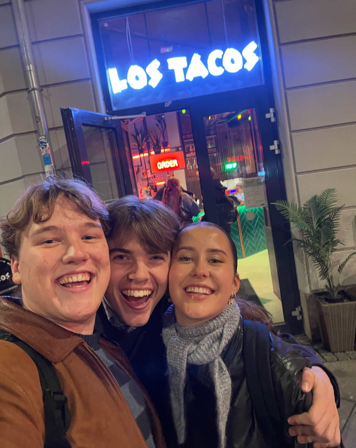 Three smiling people taking a selfie in front of a restaurant with a neon sign that says 'Los Tacos' and an interior with colorful lighting.
