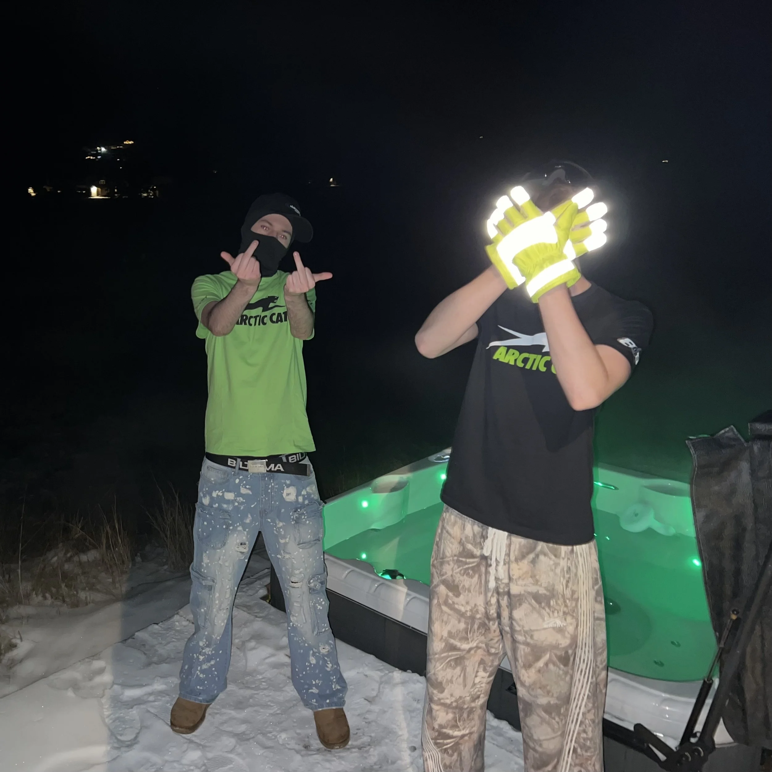 Two young men standing on snow at night near a hot tub. One is making rude hand gestures and the other, wearing reflective gloves, is holding their head, obscuring their face.