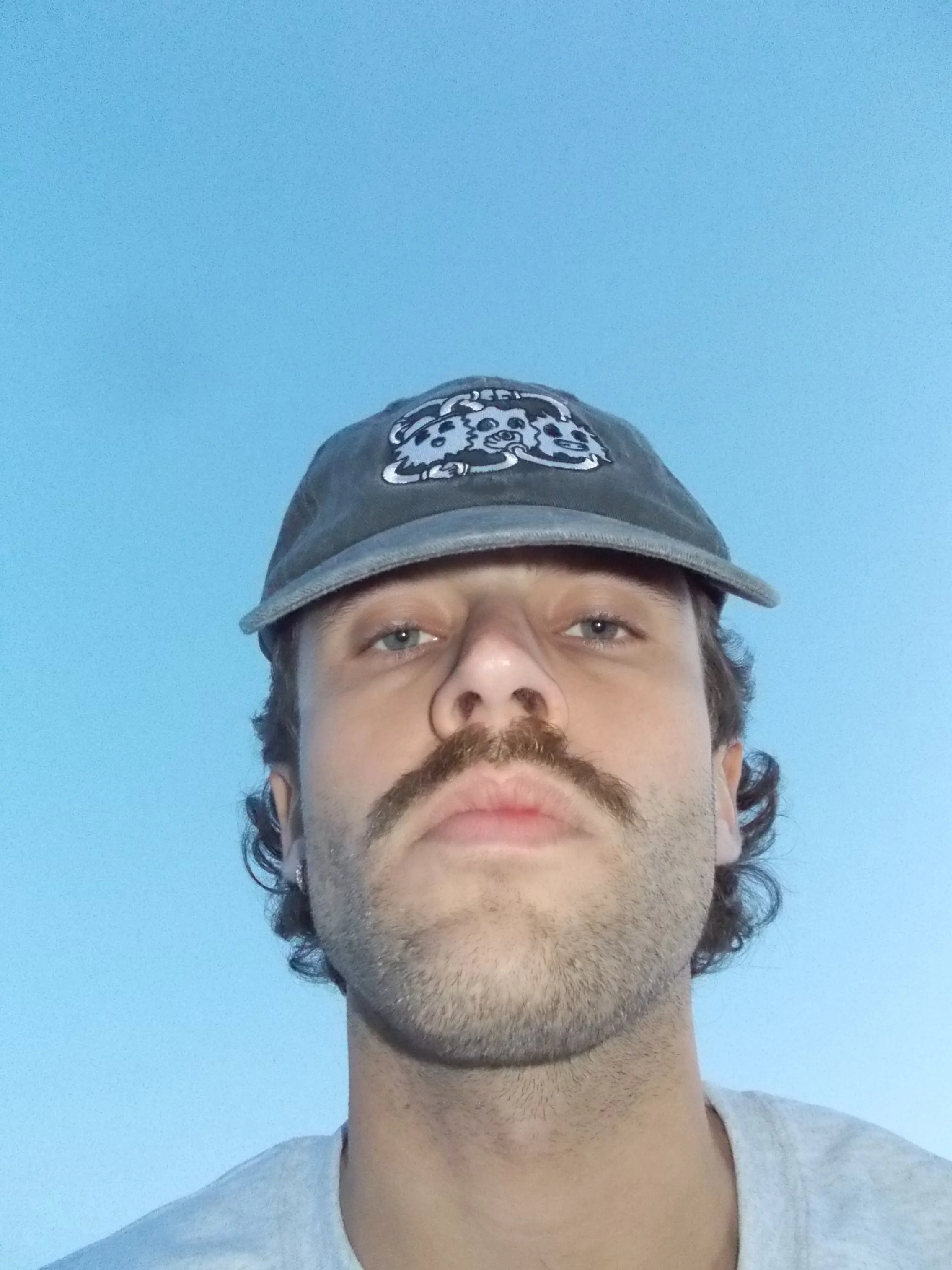 Close-up of a man with a mustache and beard, wearing a gray cap with a cartoon character on it, against a clear blue sky.