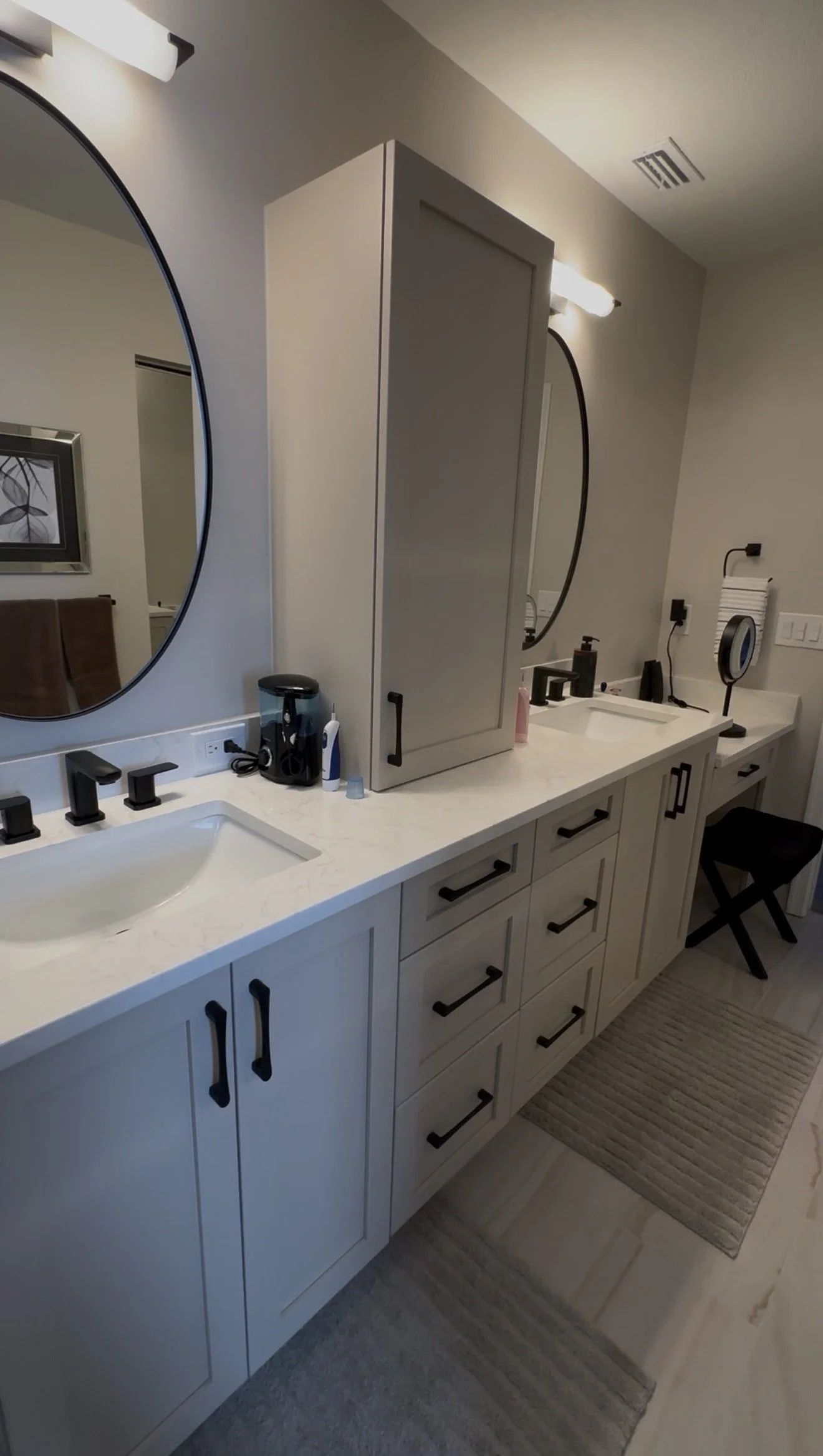 The double vanity design balances symmetry and function with the soft gray shaker cabinetry and the central countertop storage tower. The contemporary aesthetic is completed with matte black hardware, oval mirrors and LED vanity lights.