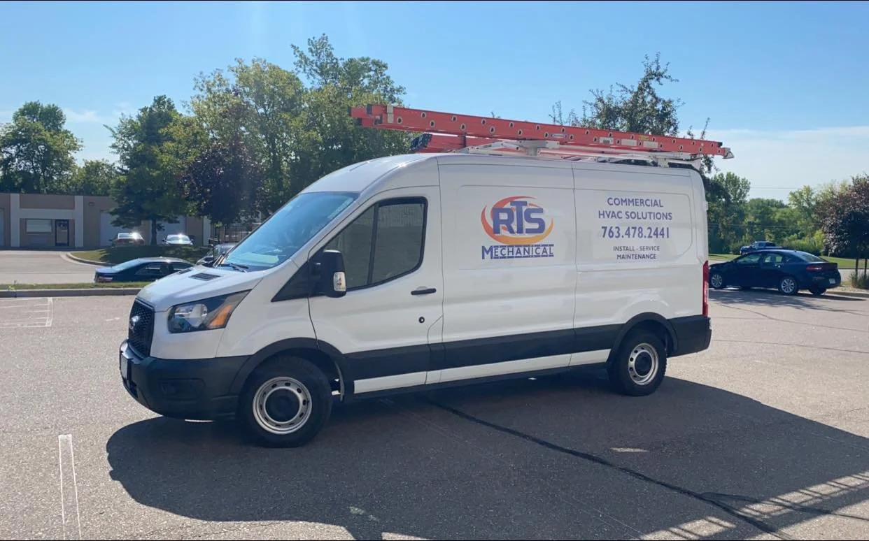 White commercial HVAC service van parked in a lot with a ladder on top, logo and contact information on the side, and trees and buildings in the background.