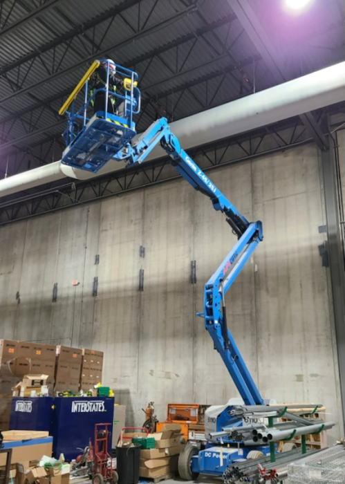 A person operating a blue boom lift inside an industrial warehouse, working on a large white pipe near the ceiling.