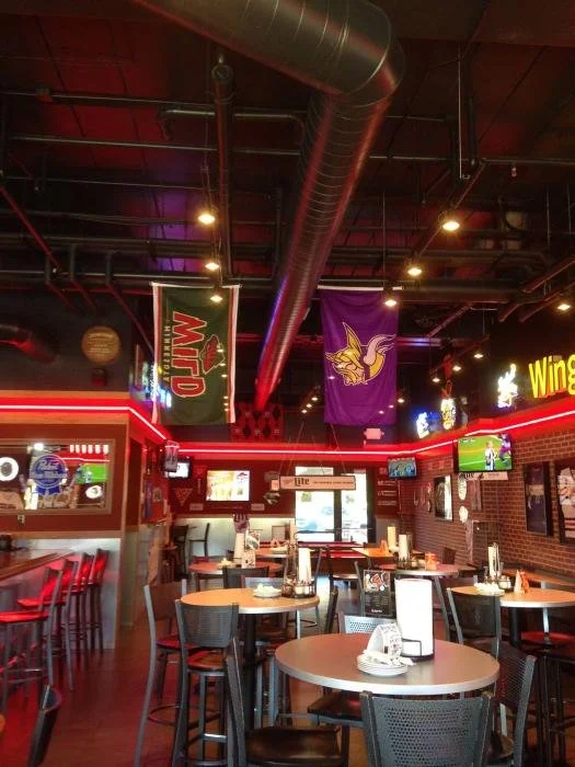 Empty sports bar with tables, chairs, and multiple TV screens showing sports games, decorated with flags and sports banners.