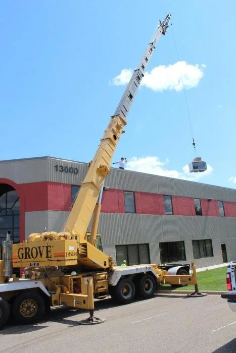 A large yellow crane lifting a small building module in front of a multi-story commercial building with a gray and red facade.
