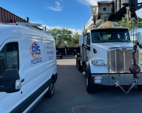 White utility truck with a bucket lift parked next to a white service van with logo and text, in a parking lot with trees and blue sky in the background.