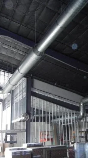 Industrial interior with large metal ventilation pipe and metal bars in the foreground, ceiling with black insulation and exposed ductwork.