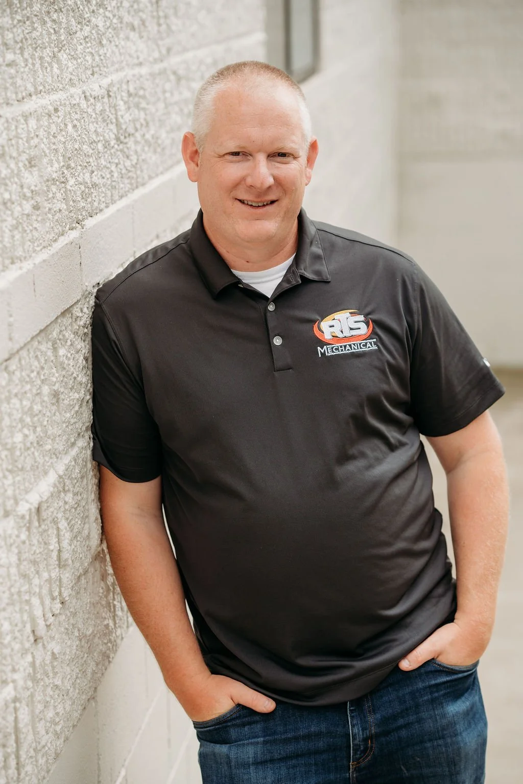 A man with short hair, wearing a black polo shirt with a logo, standing next to a textured concrete wall, smiling at the camera.