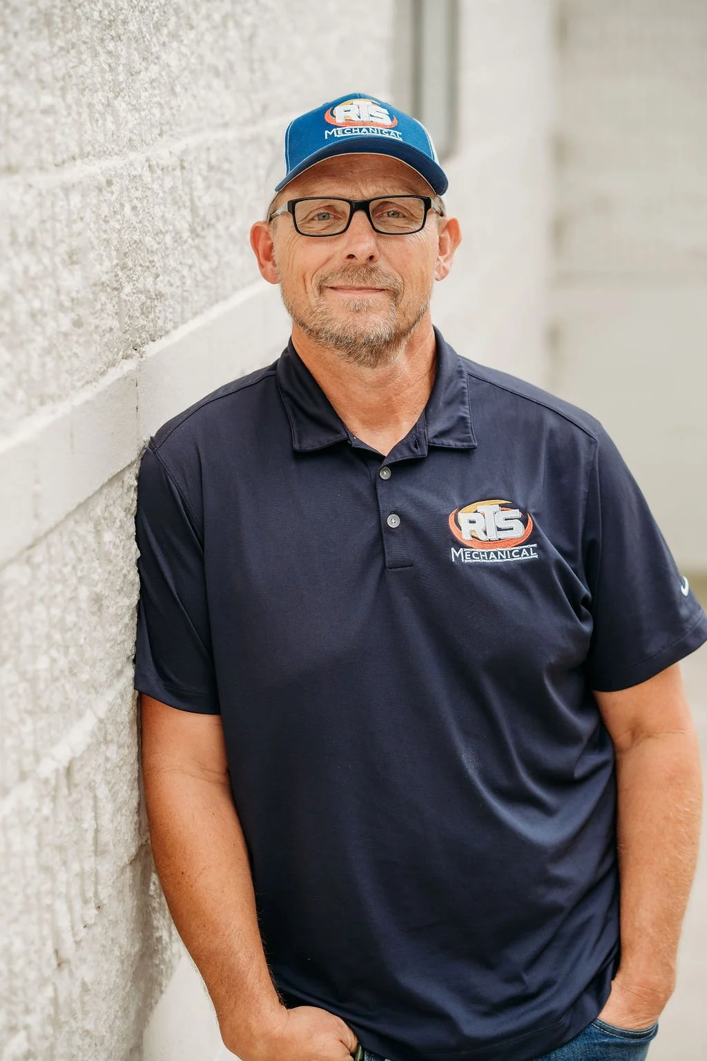 A middle-aged man wearing glasses, a navy blue polo shirt, and a matching cap with the 'RIS Mechanical' logo, standing against a textured concrete wall with one hand in his pocket.