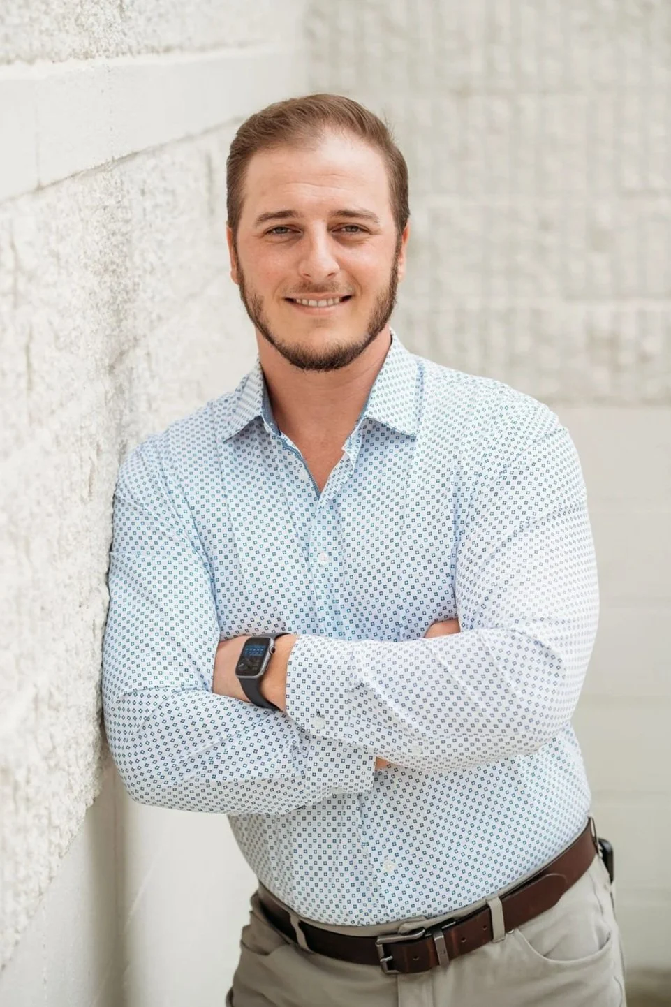 A man with light brown hair and a beard poses with arms crossed, leaning against a white brick wall, wearing a light blue patterned shirt and a black smartwatch.