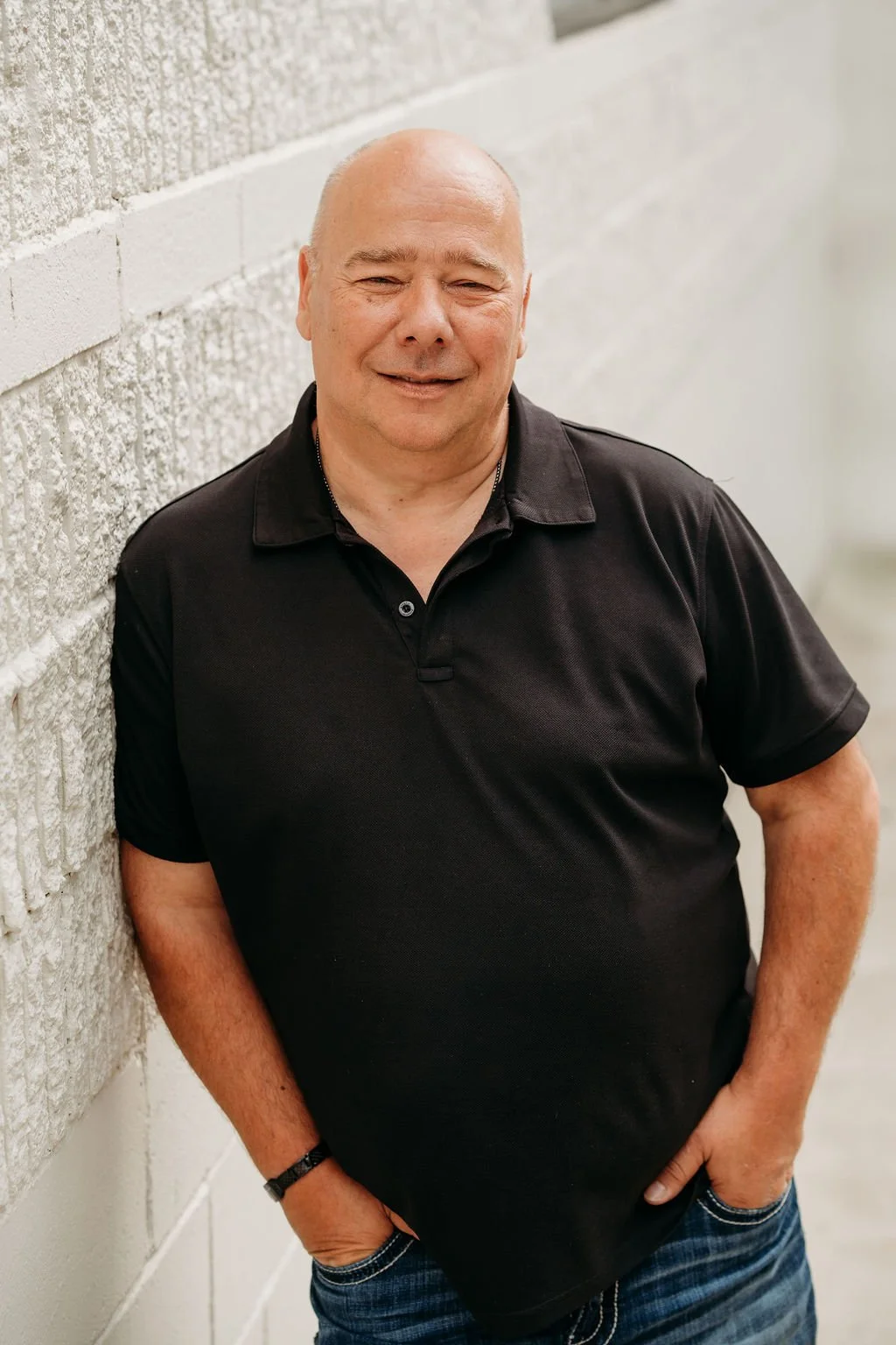 A middle-aged man with a bald head leaning against a textured concrete wall, smiling, wearing a black polo shirt and blue jeans, with hands in pockets.