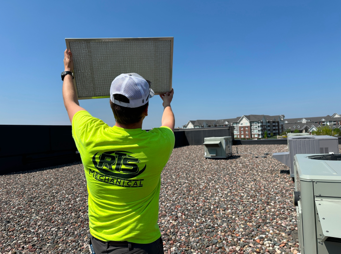 A person wearing a neon yellow shirt and a white cap installs new commercial filter on a building rooftop.
