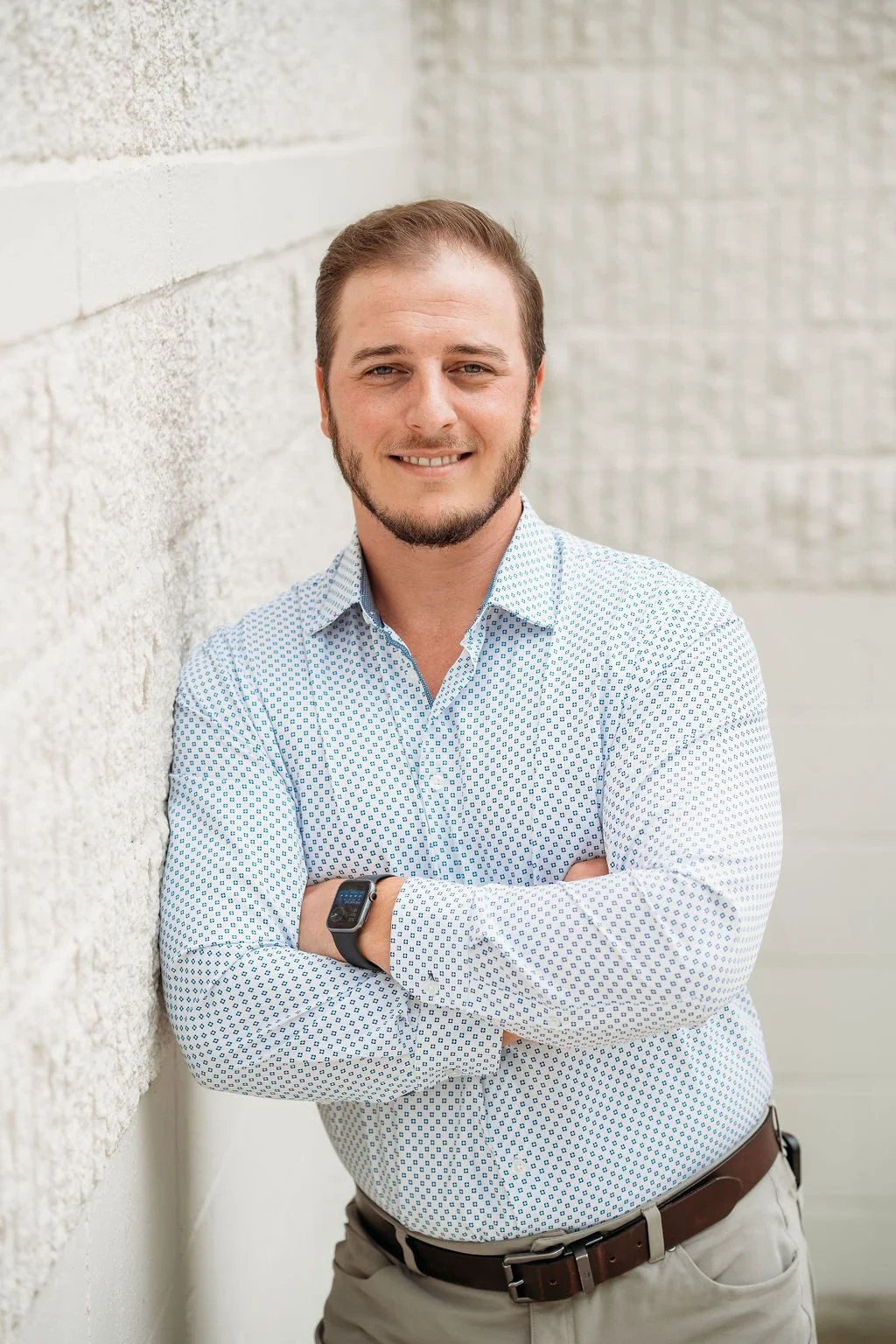 A young man with light brown hair, beard, and blue eyes smiling while standing with arms crossed against a textured beige wall, wearing a white patterned button-up shirt and a smartwatch.