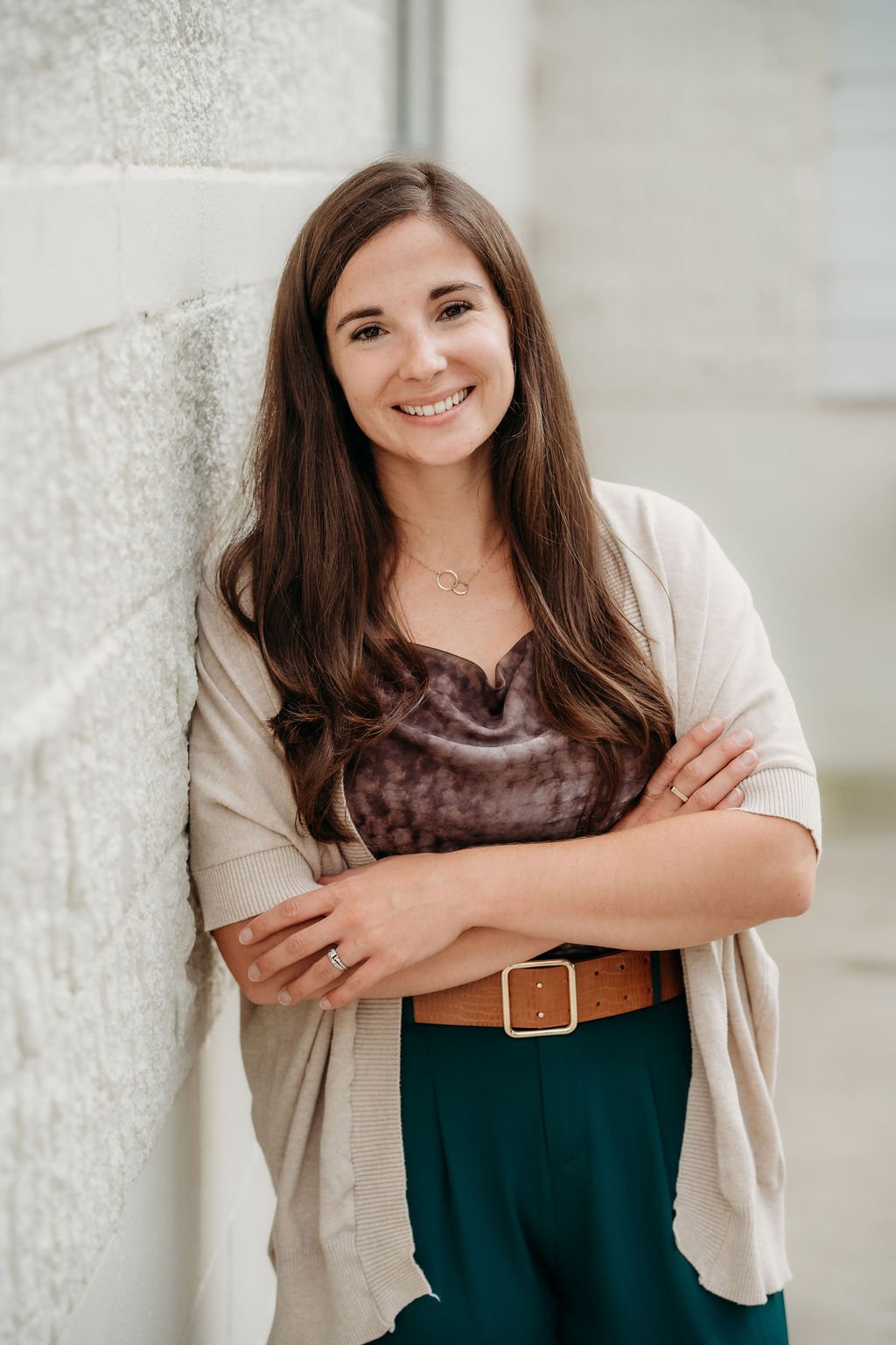 A smiling woman with long brown hair leaning against a light-colored brick wall with her arms crossed, wearing a beige cardigan, a patterned blouse, and teal pants.