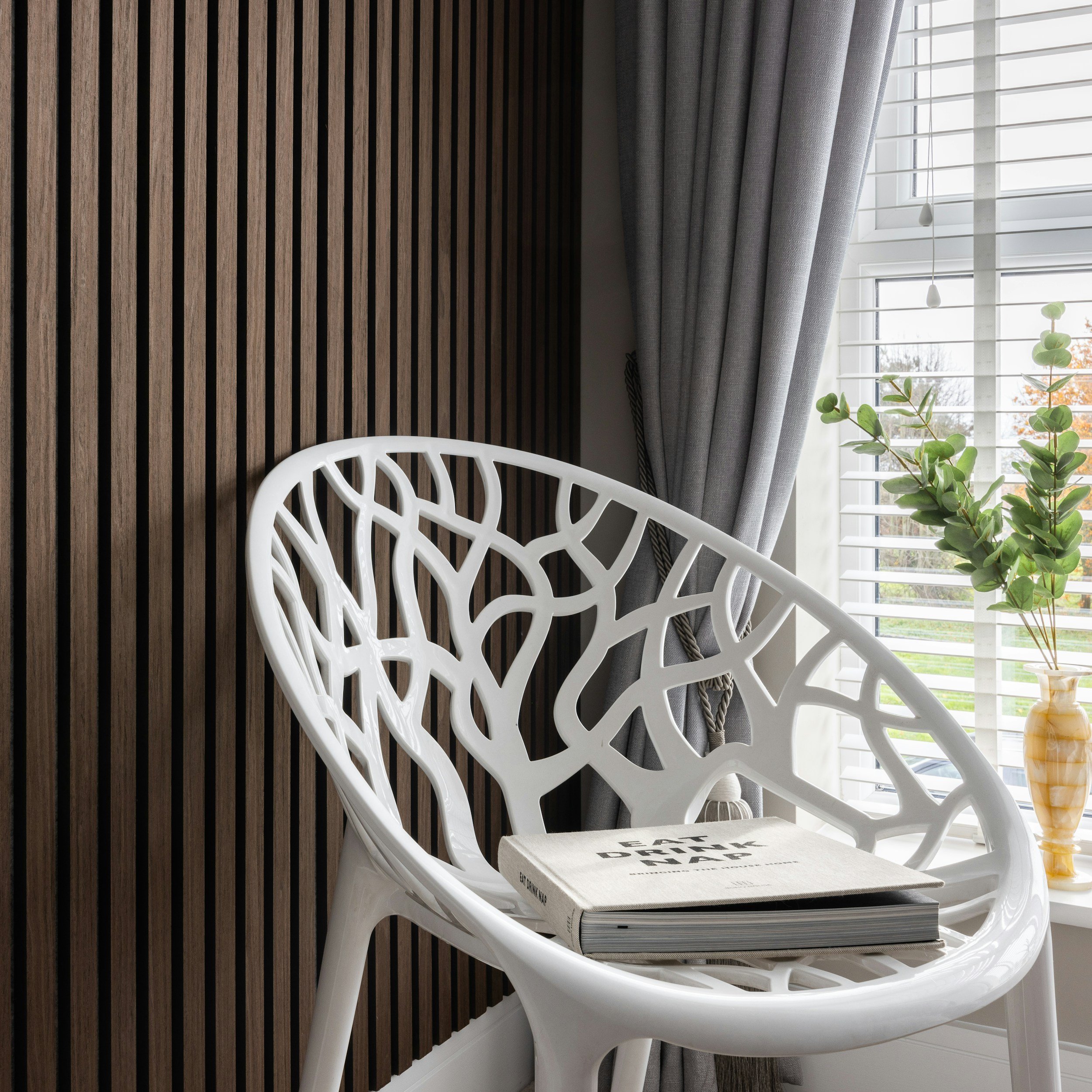 Modern indoor corner with a white decorative chair with a tree branch pattern, a beige book on the seat, dark wood slatted wall, gray curtains, and a window with horizontal blinds and a potted plant.