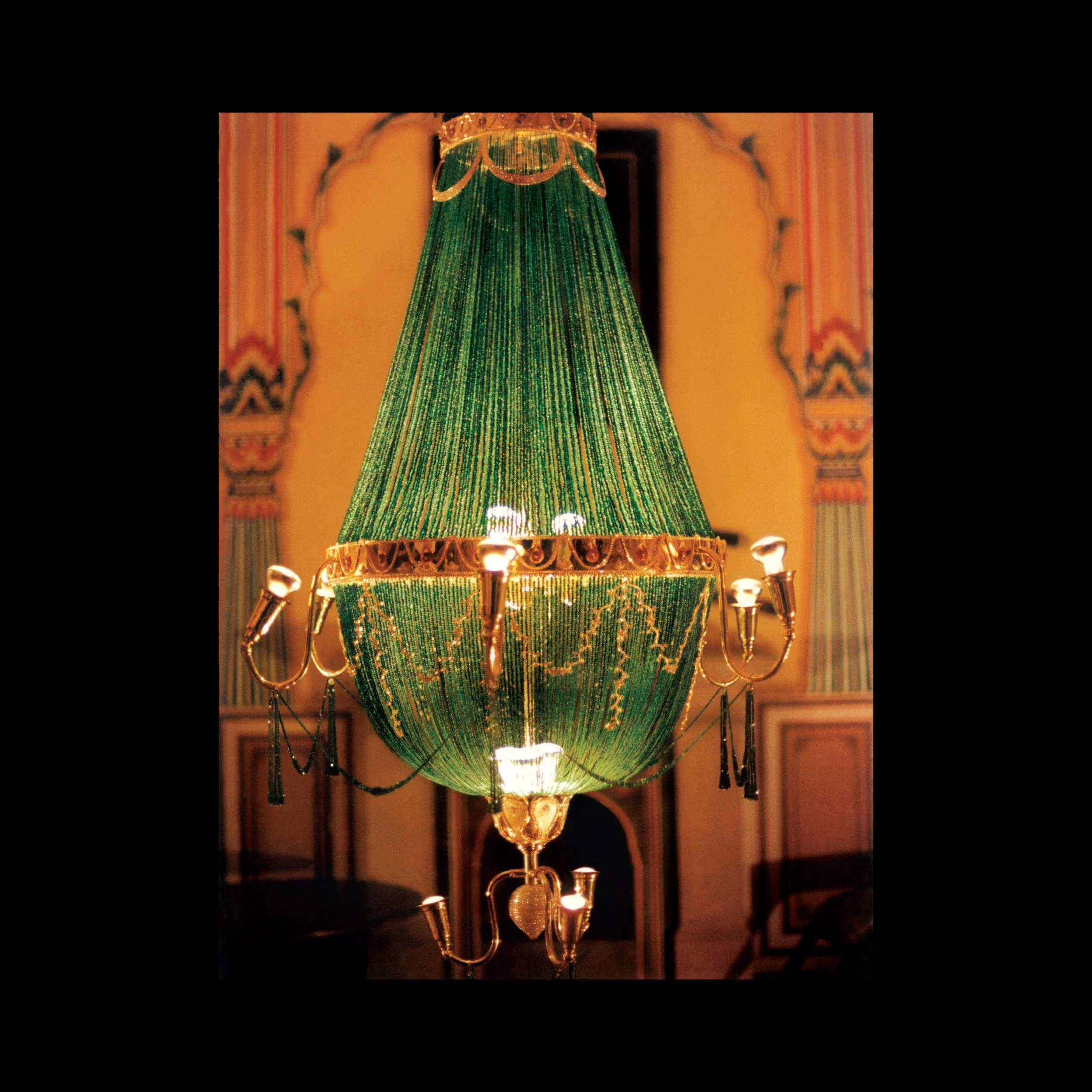 An ornate chandelier with green beaded lampshade and gold accents, hanging in a decorated room with patterned walls.