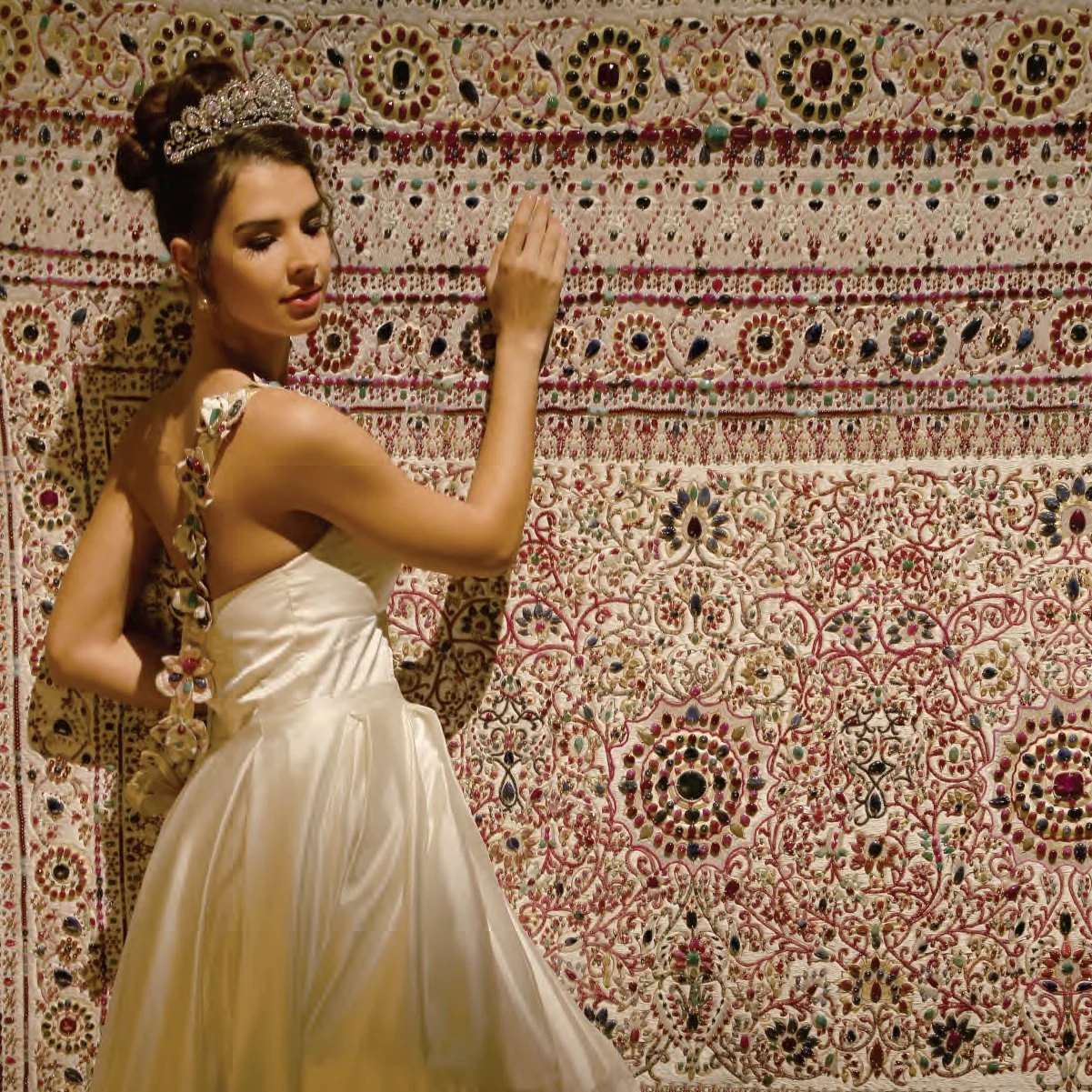 A woman in an elegant, cream-colored gown with intricate accessories, standing against a richly decorated, embroidered wall with patterns and embellishments.