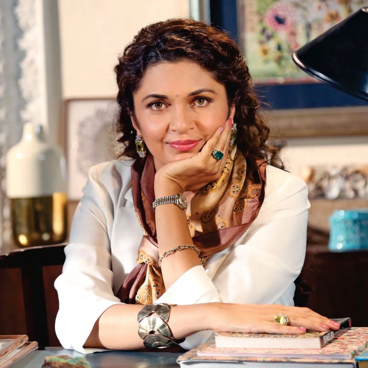 A woman with dark, curly hair and light brown eyes, wearing jewelry including earrings, rings, a bracelet, and a watch, sitting at a desk with her chin resting on her hand, smiling slightly at the camera in a cozy room with artwork and decorative items in the background.