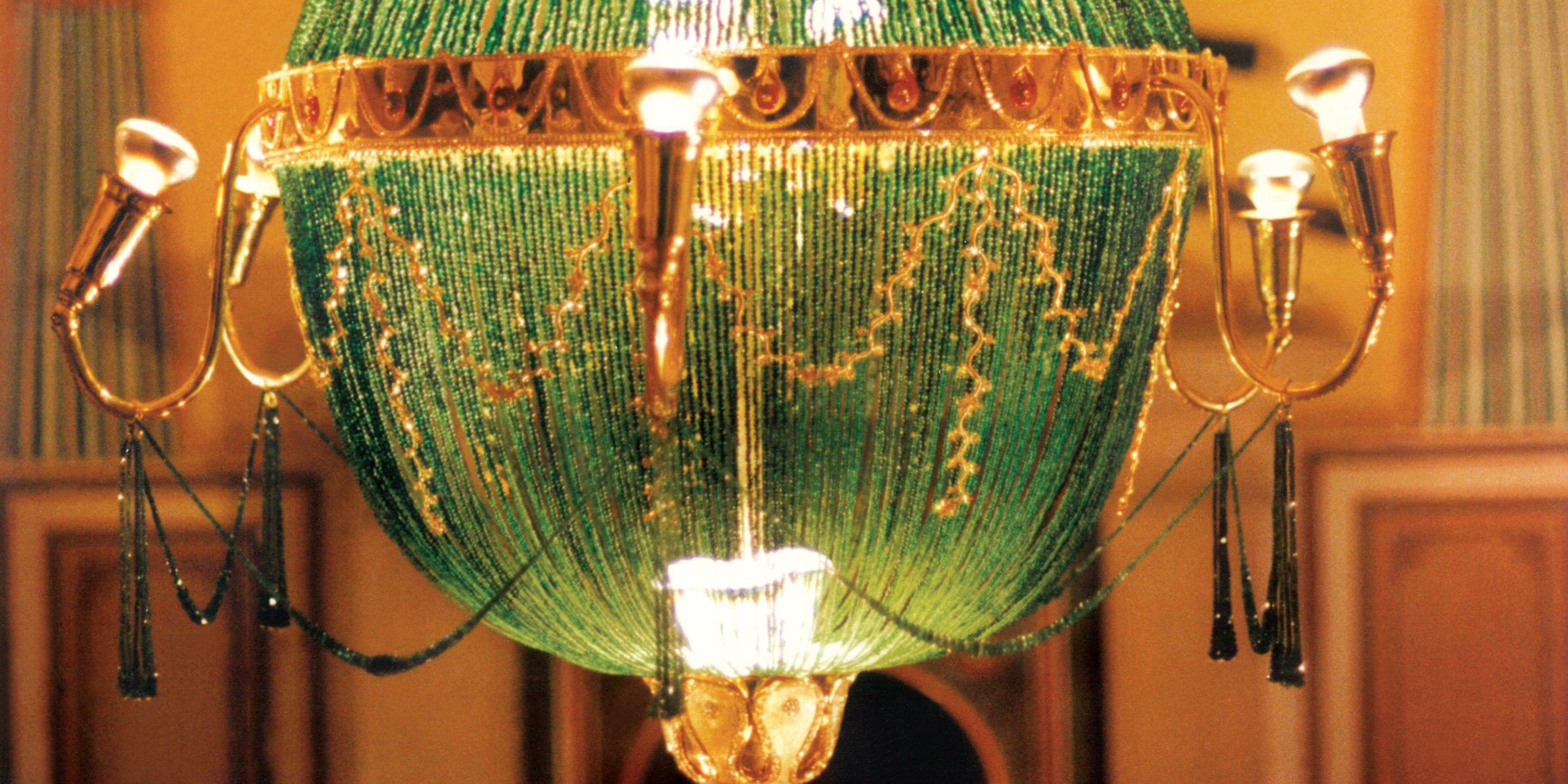 A close-up view of an ornate chandelier with green beaded design, gold accents, and hanging chains, mounted in an elegant room with patterned wallpaper.