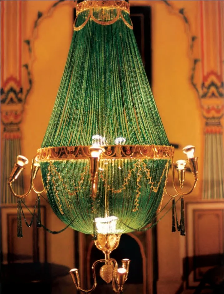 A large ornate chandelier with a green beaded lampshade and gold accents, hanging in an elegant room with decorative wall moldings.