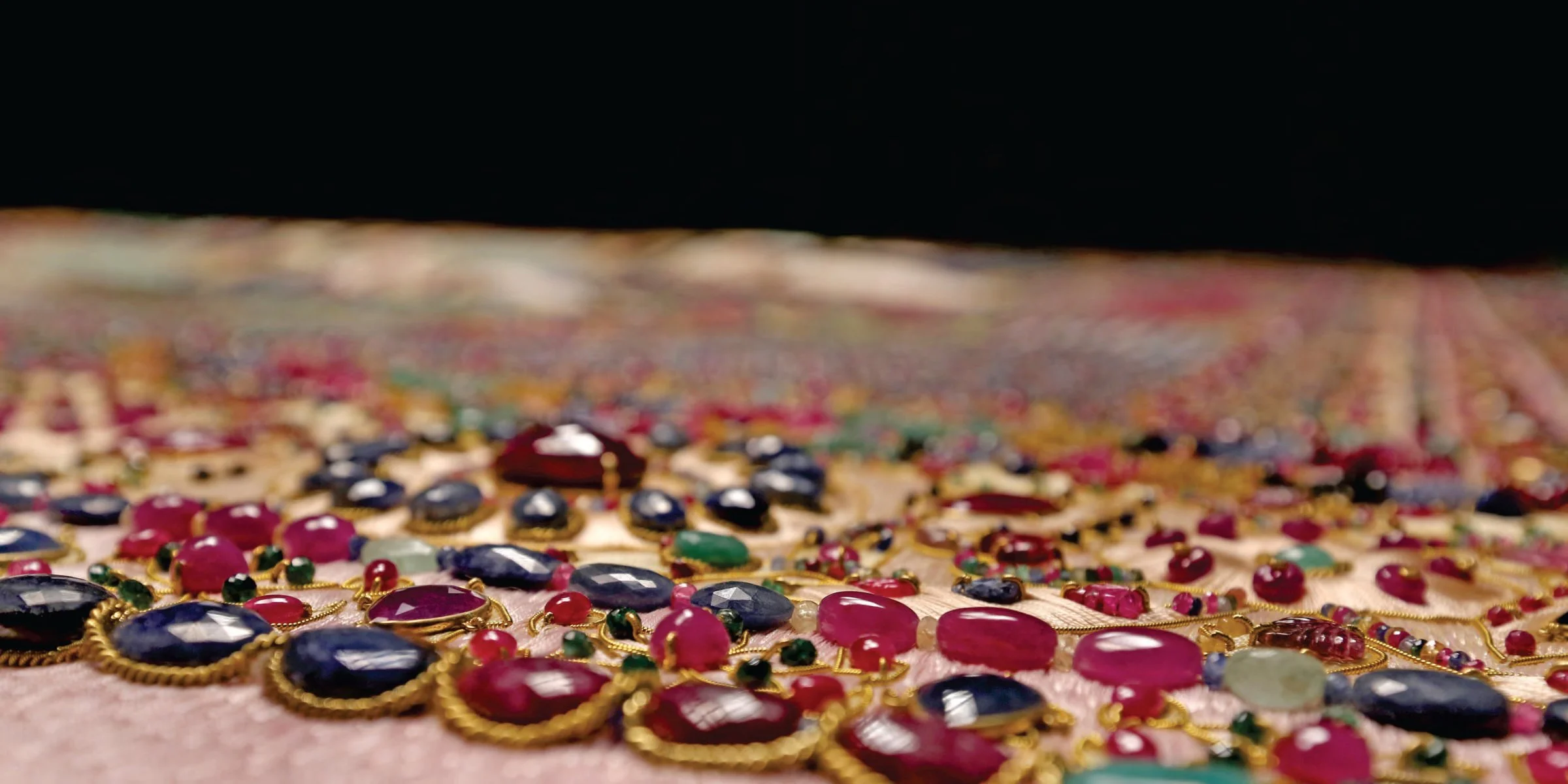 Close-up of scattered colorful gemstone jewelry, including red, blue, green, and black stones with gold settings, on a fabric surface with a blurred colorful background.