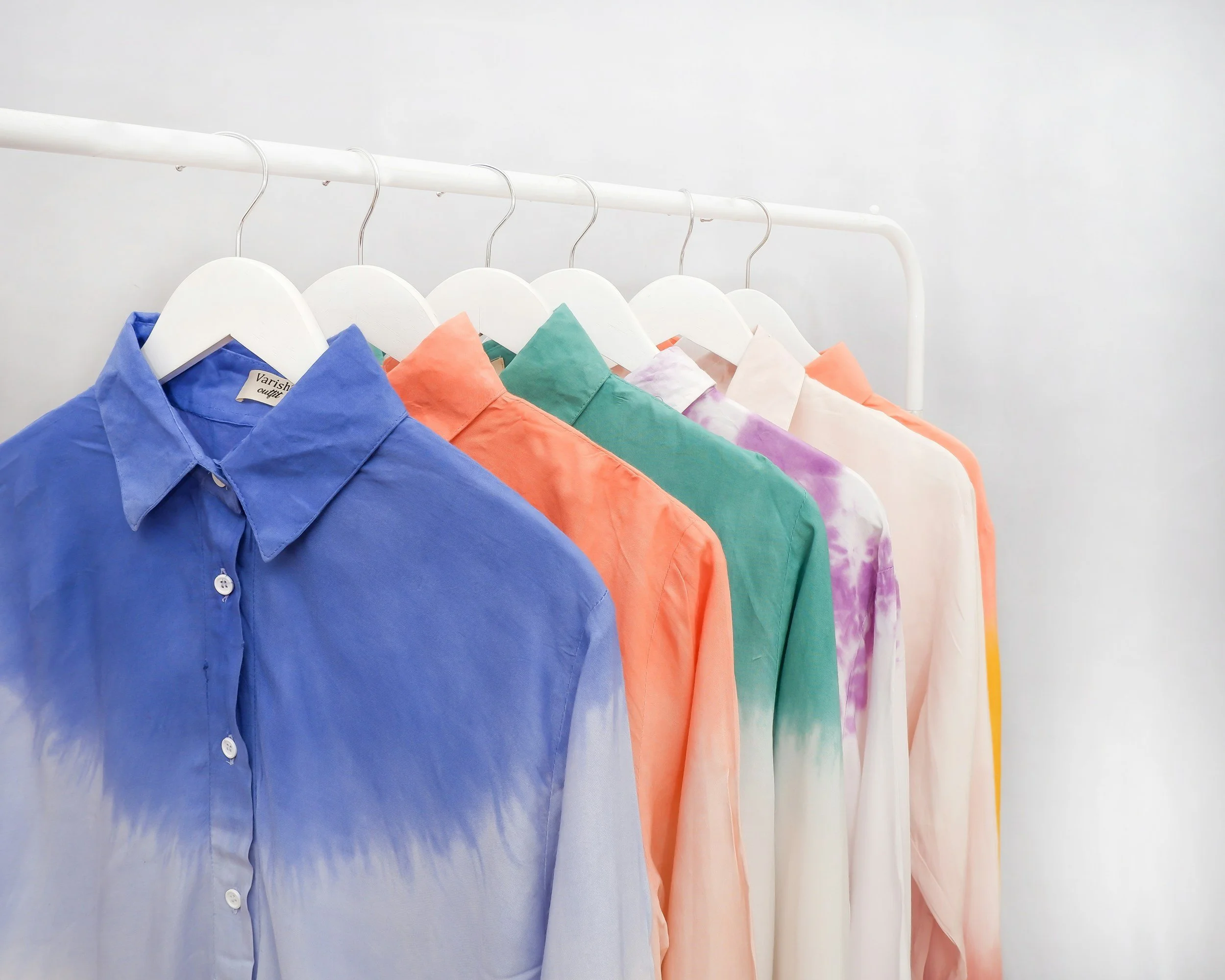 Six colorful collared shirts hanging on a white clothing rack against a plain white wall.